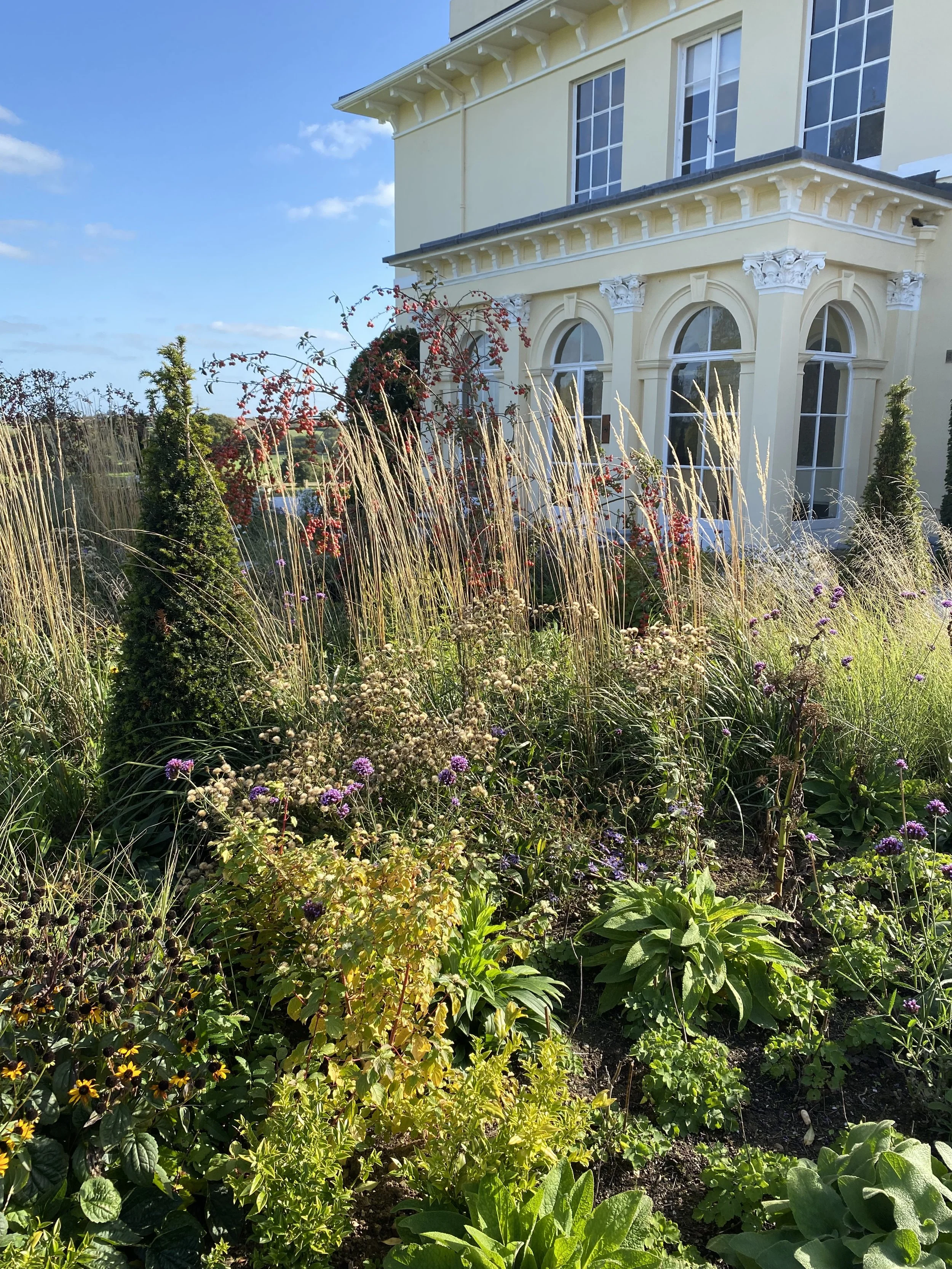 A garden in front of a large, pale yellow house with decorative white trim and tall, arched windows. The garden has a variety of plants, including purple, yellow, and green foliage, with tall ornamental grasses and flowering shrubs, under a clear blue sky.