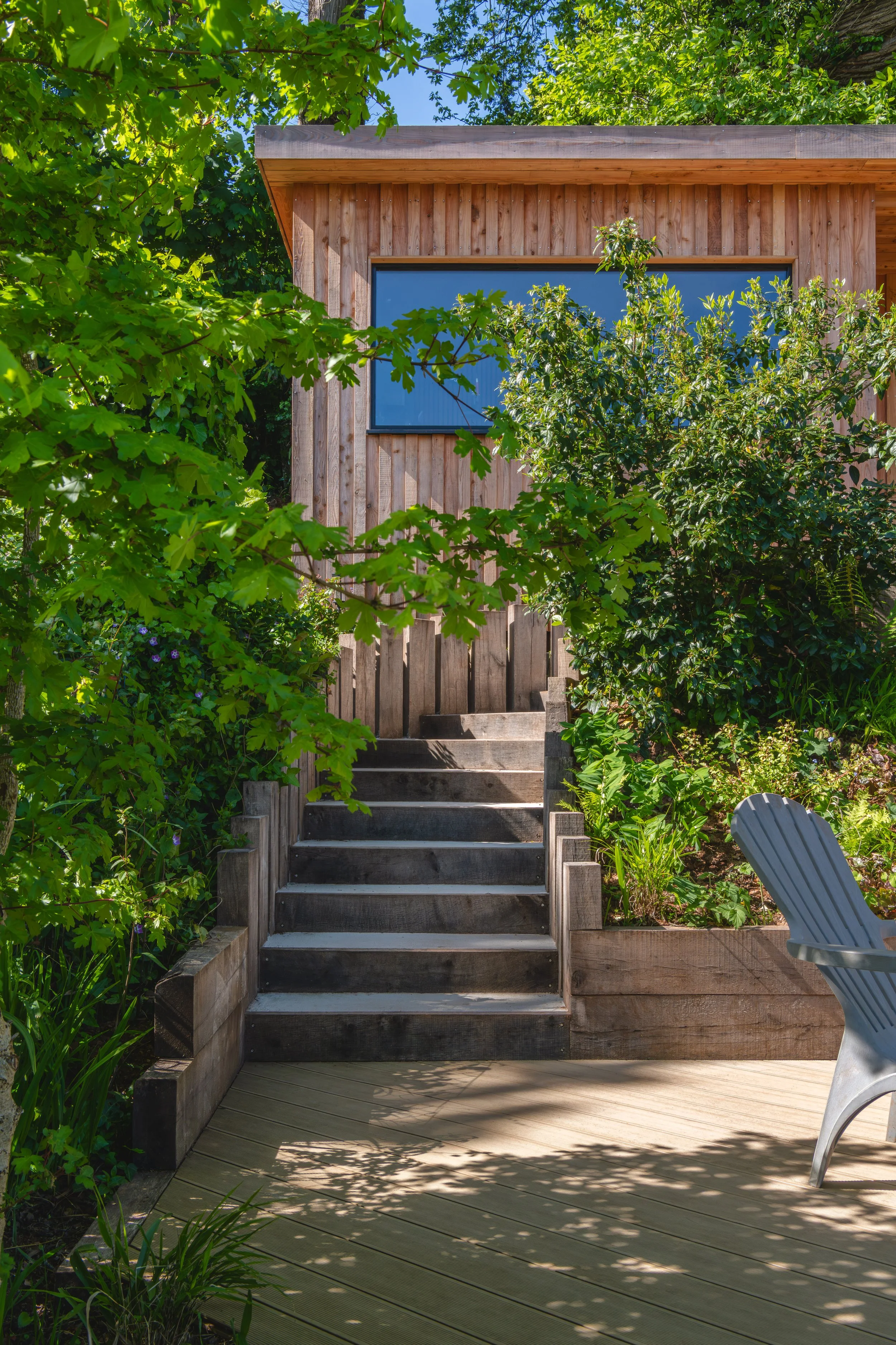 Wooden steps leading up to a garden office with a large window, surrounded by green foliage and sunlight filtering through the trees, with a chair on a wooden deck.