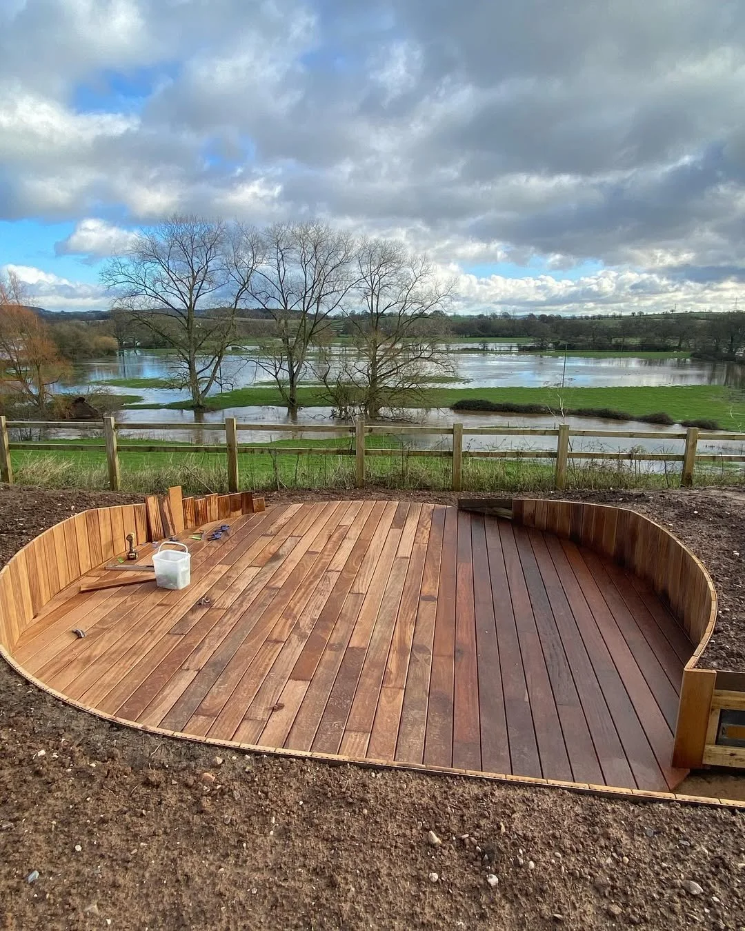A newly constructed wooden deck with a curved design, set on bare soil, overlooking a scenic landscape with a river, trees, and cloudy sky.