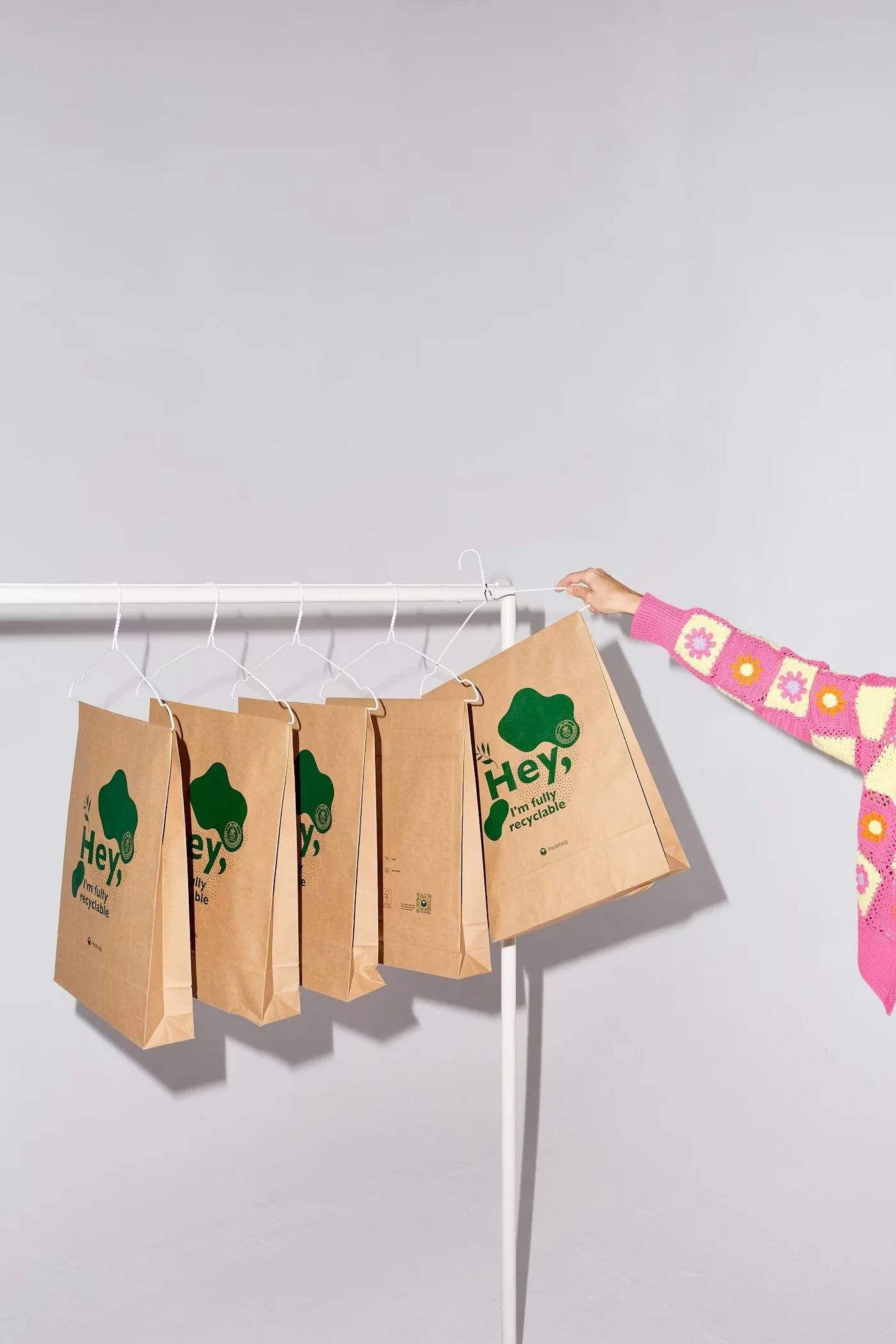 A person is hanging paper shopping bags on a white clothing rack. The bags are brown with green printed rescue logos and text that says 'Hey, I'm fully recyclable.' The person's arm is visible, dressed in a colorful knitted sweater with pink, white, and yellow flower patterns. The background is a plain light gray wall.
