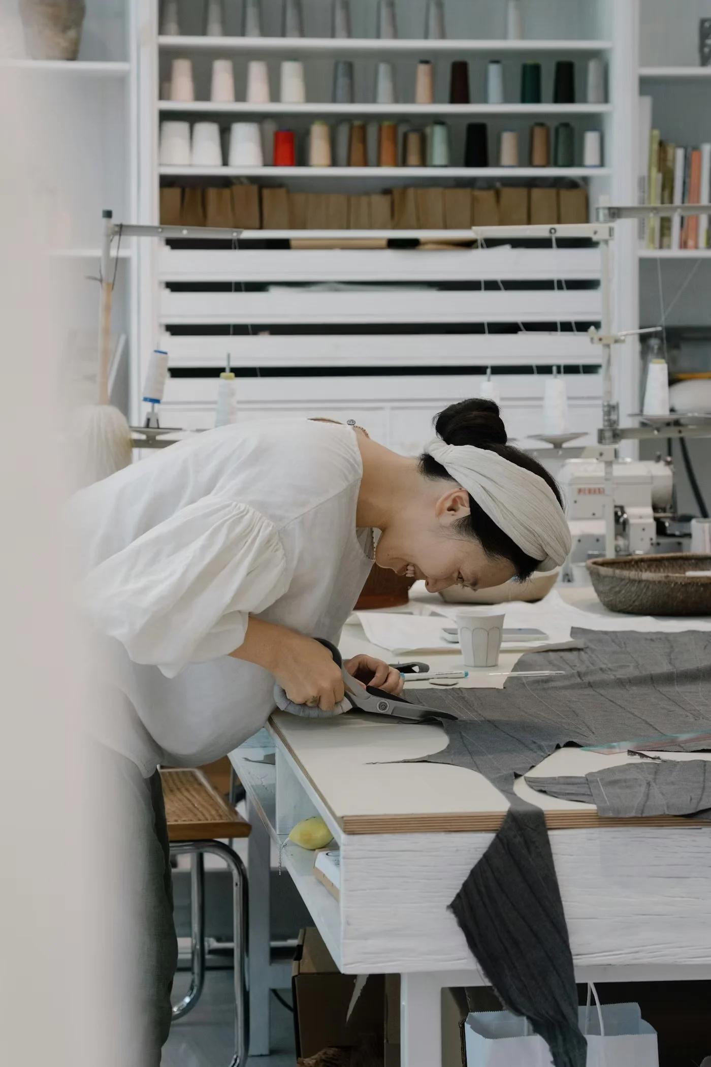 A woman with a headband is smiling and working on fabric with scissors at a sewing or textile workspace.