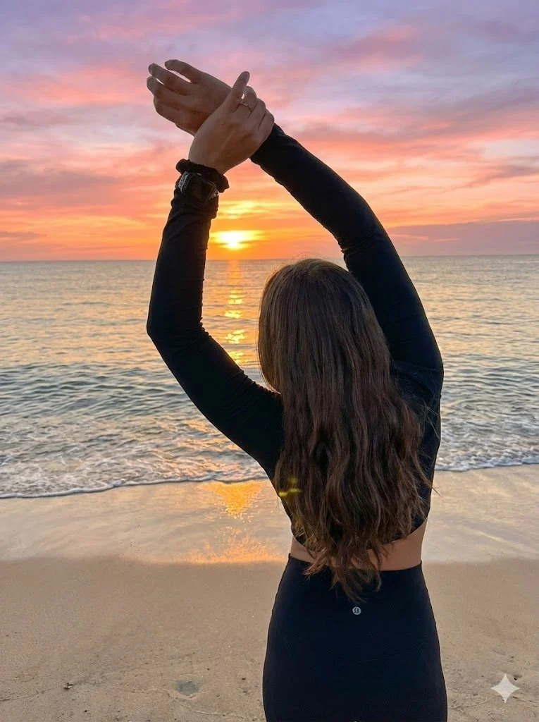 Person practicing yoga or stretching on the beach at sunset, with the ocean and colorful sky in the background.