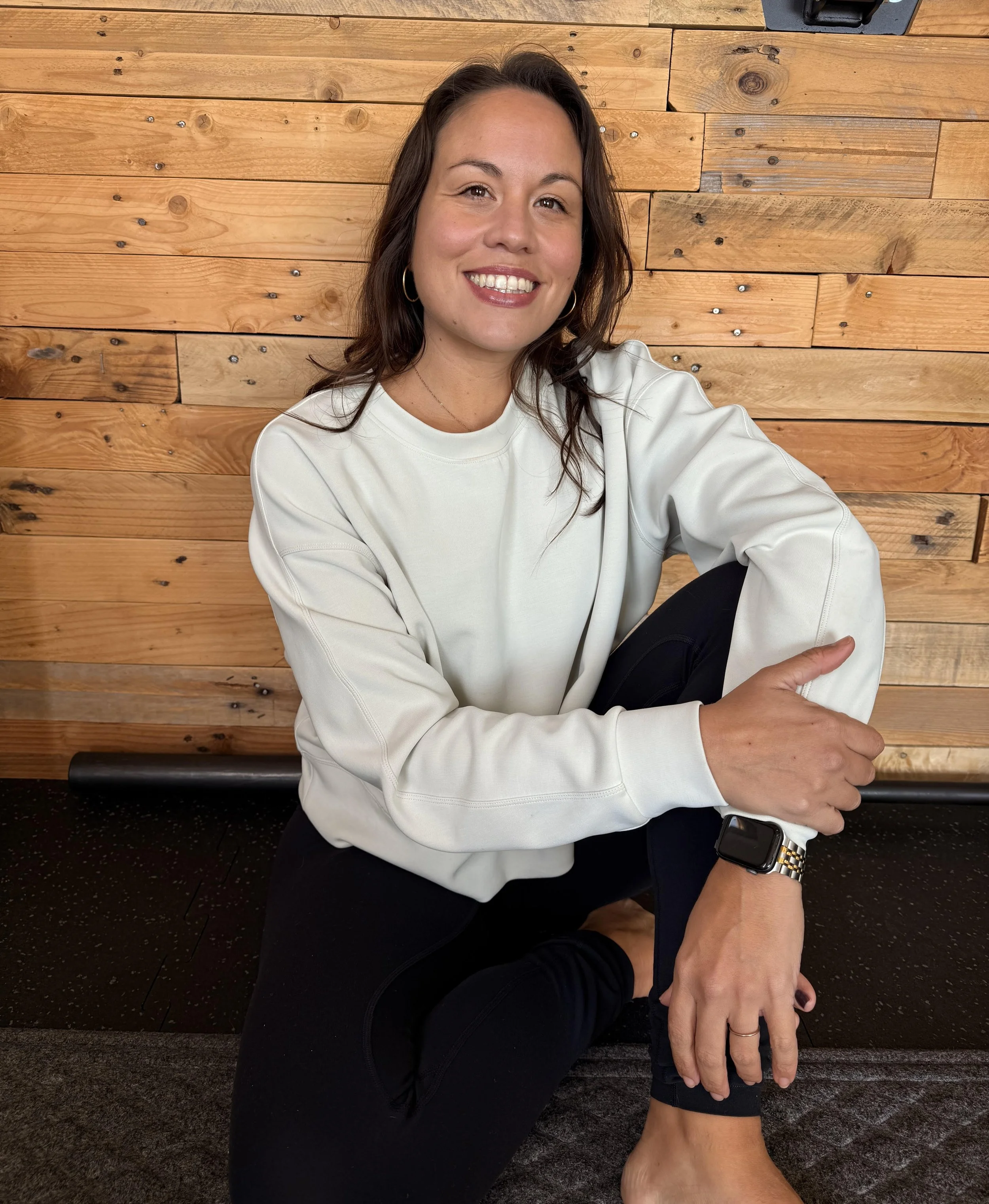 A woman smiling, sitting on a gym floor with a wooden wall in the background, wearing a white sweatshirt, black leggings, and a smartwatch.
