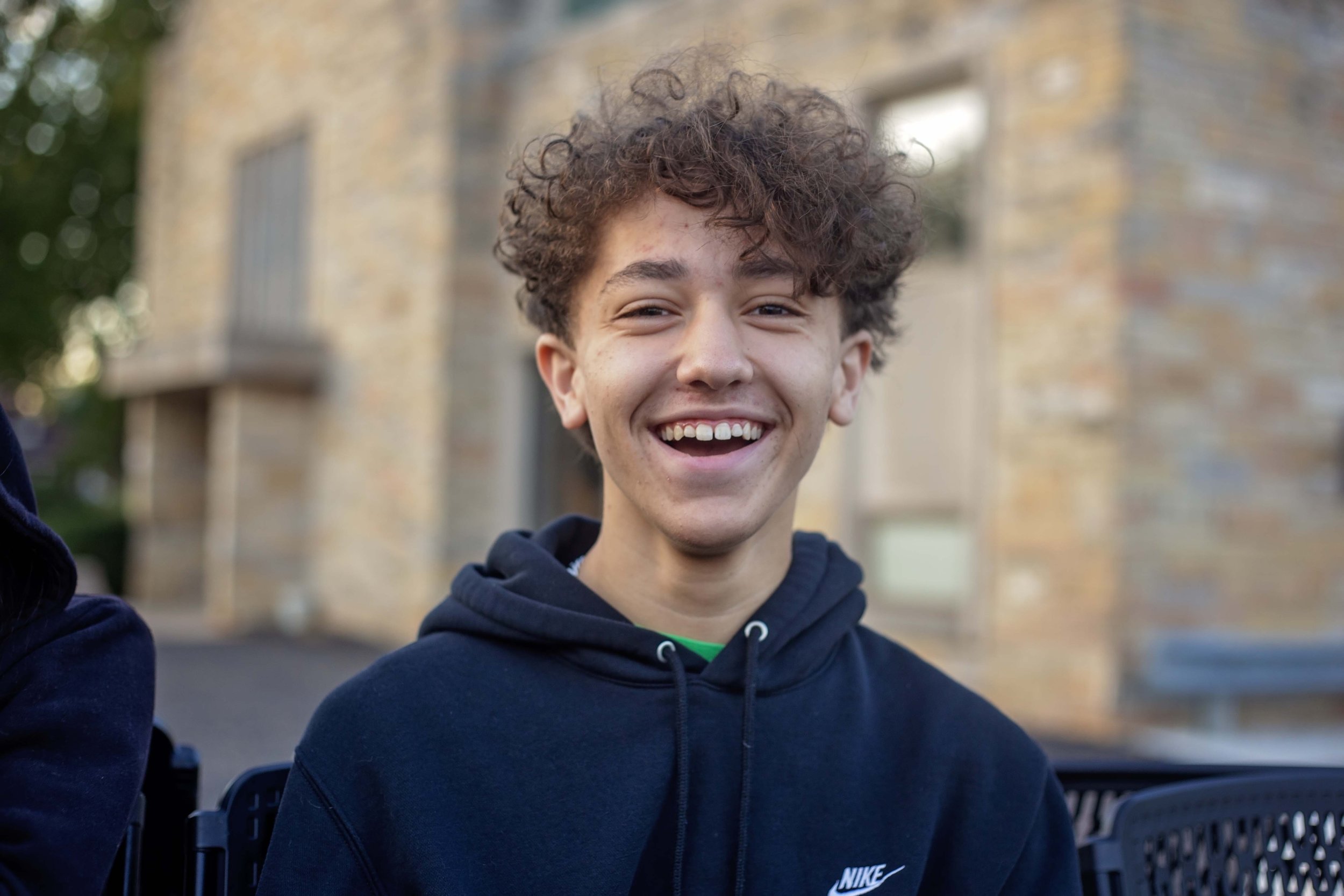 A young man with curly brown hair smiling outdoors with a blurred brick building in the background, wearing a black Nike hoodie.
