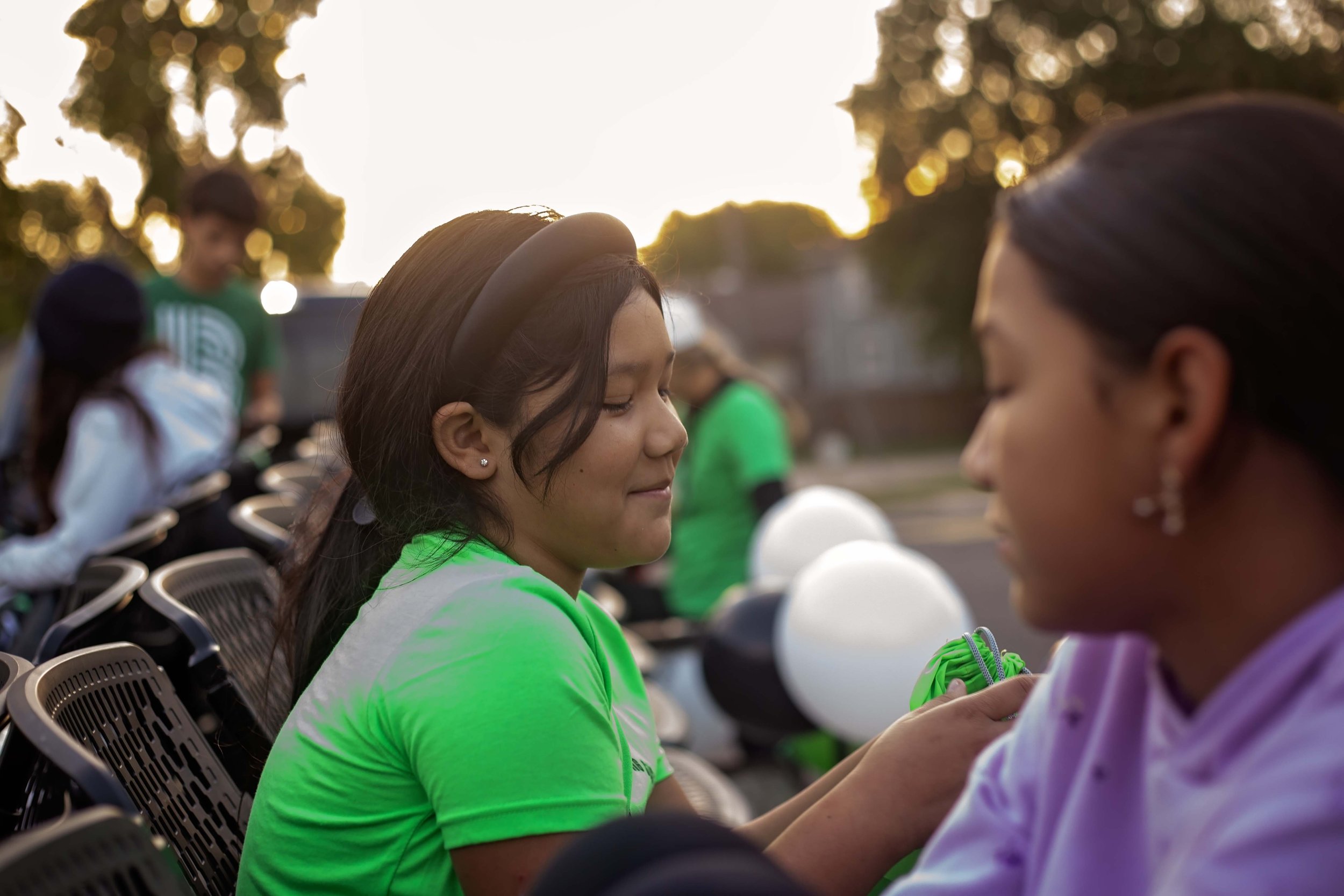 A woman with dark hair, wearing a black headband and a neon green shirt, prays with her eyes closed outdoors in the late afternoon or early evening sunlight. She is sitting next to another woman with dark hair pulled back, also with her eyes closed. There are chairs and people, some holding white balloons, in the background.