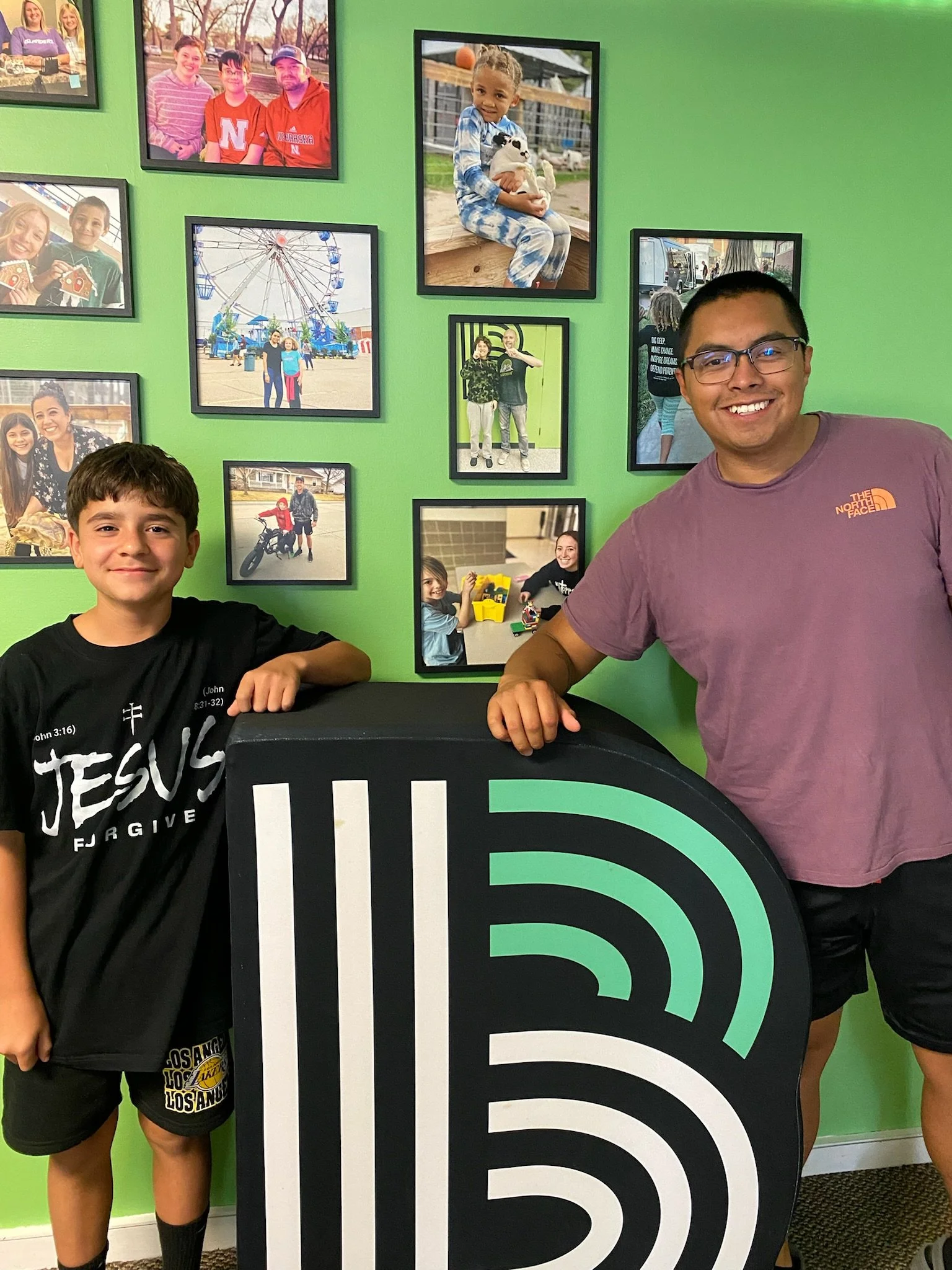 Two boys are standing indoors next to a large black sign with colorful Wi-Fi signal icon, in front of a green wall decorated with framed family photos.