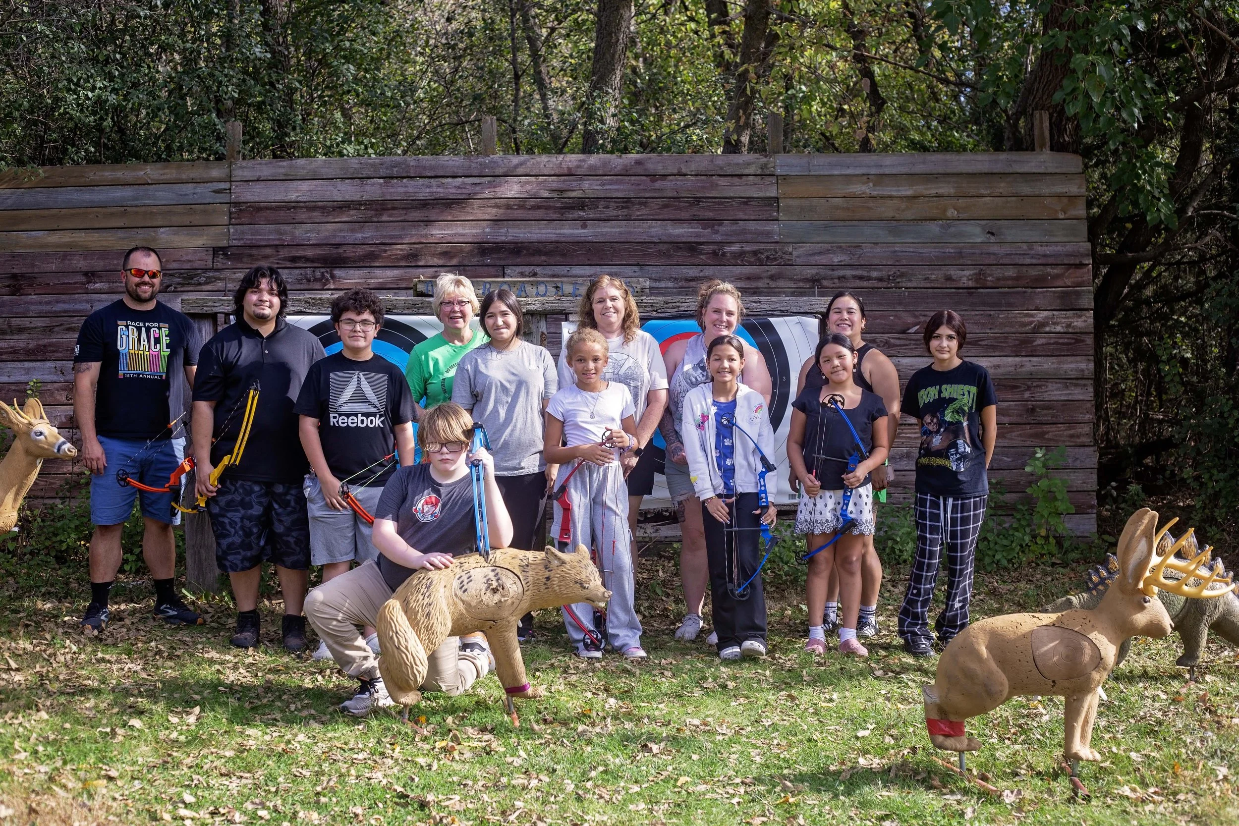 Group of people, including children and adults, standing outdoors in front of a wooden wall with trees in background. Some children are holding archery bows and arrows. There are three animal-shaped targets and leaf-covered ground.
