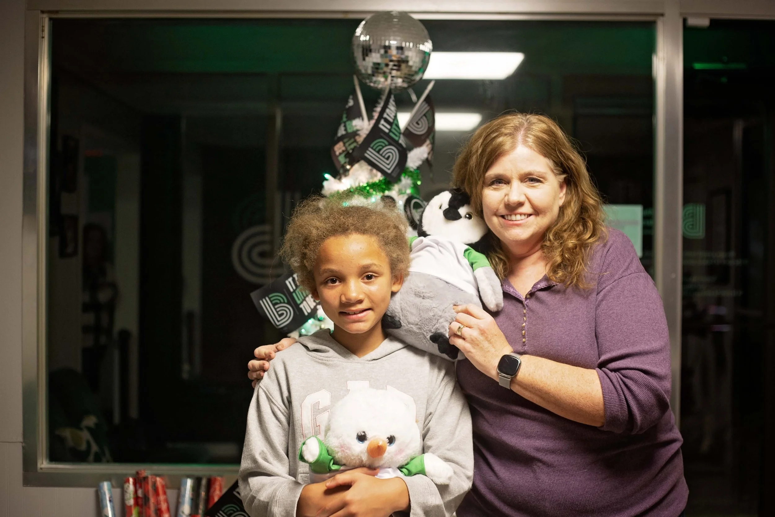 A woman and a young girl smiling and holding plush toys in front of a decorated window with green lights, balloons, and the number 50. The woman has curly red hair and is wearing a purple sweater. The girl has curly hair and is wearing a gray hoodie.