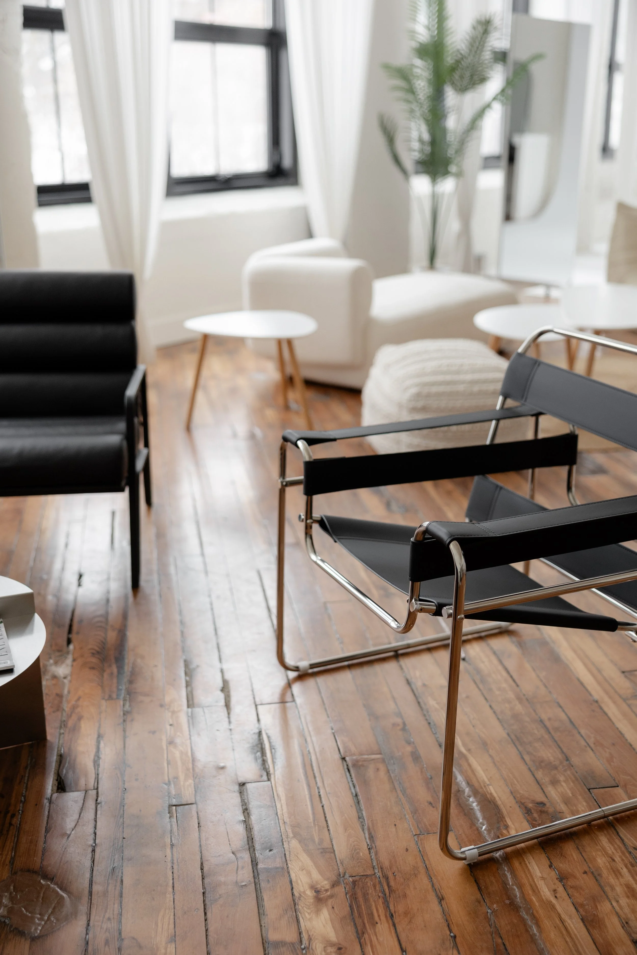 Modern living room with black and gray chairs, white side tables, a white armchair, and a large window with white curtains.