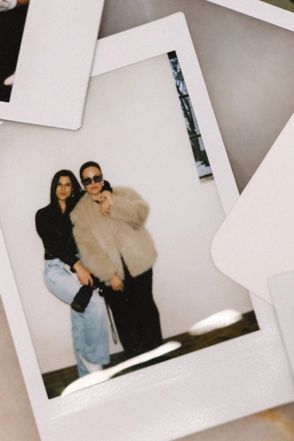 A photograph of two women, one wearing sunglasses and a fur coat, the other in a black jacket and light blue jeans, posing together indoors.