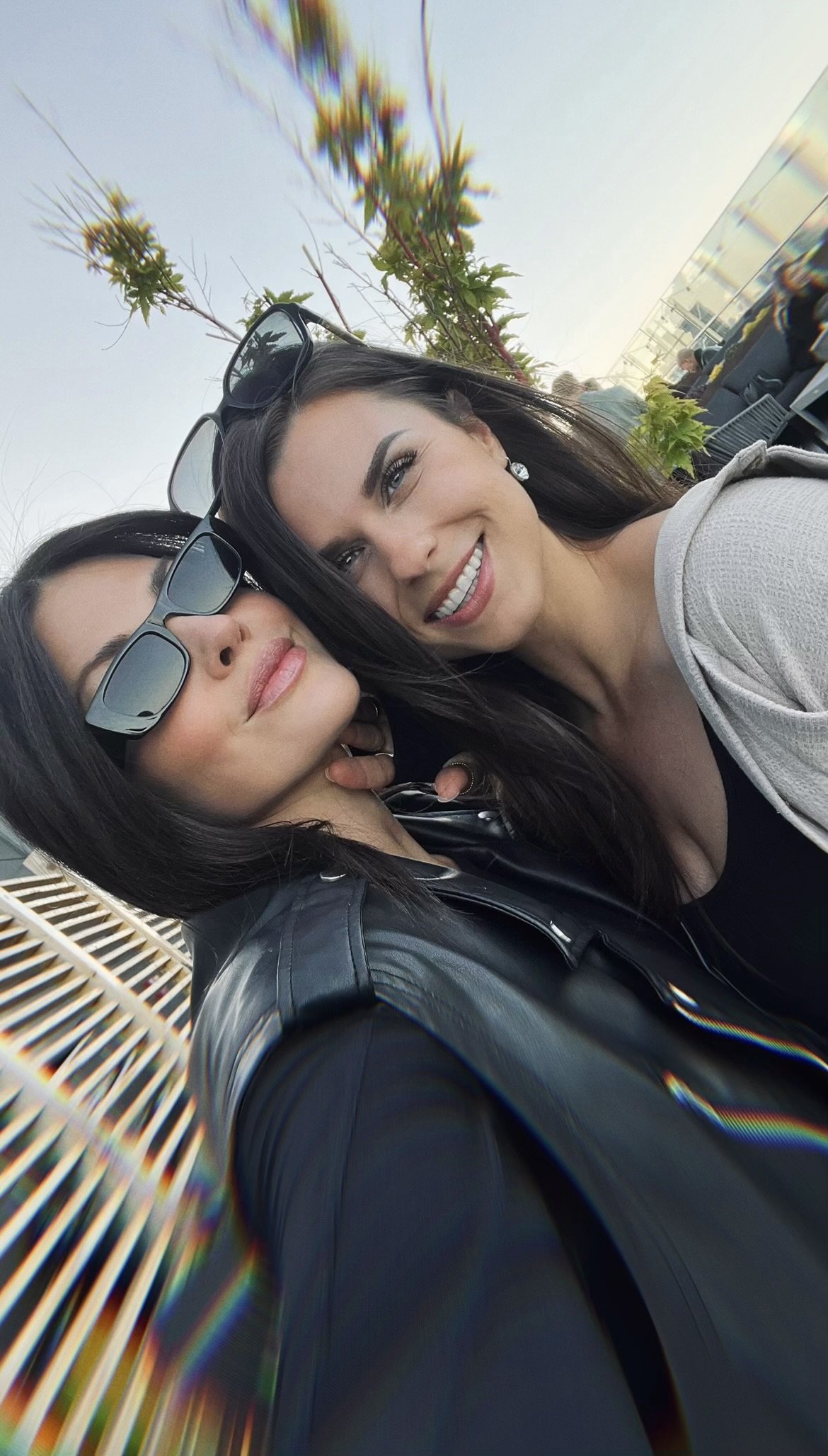 Two women taking a selfie outdoors, one with sunglasses and the other smiling, with a tree and people in the background.