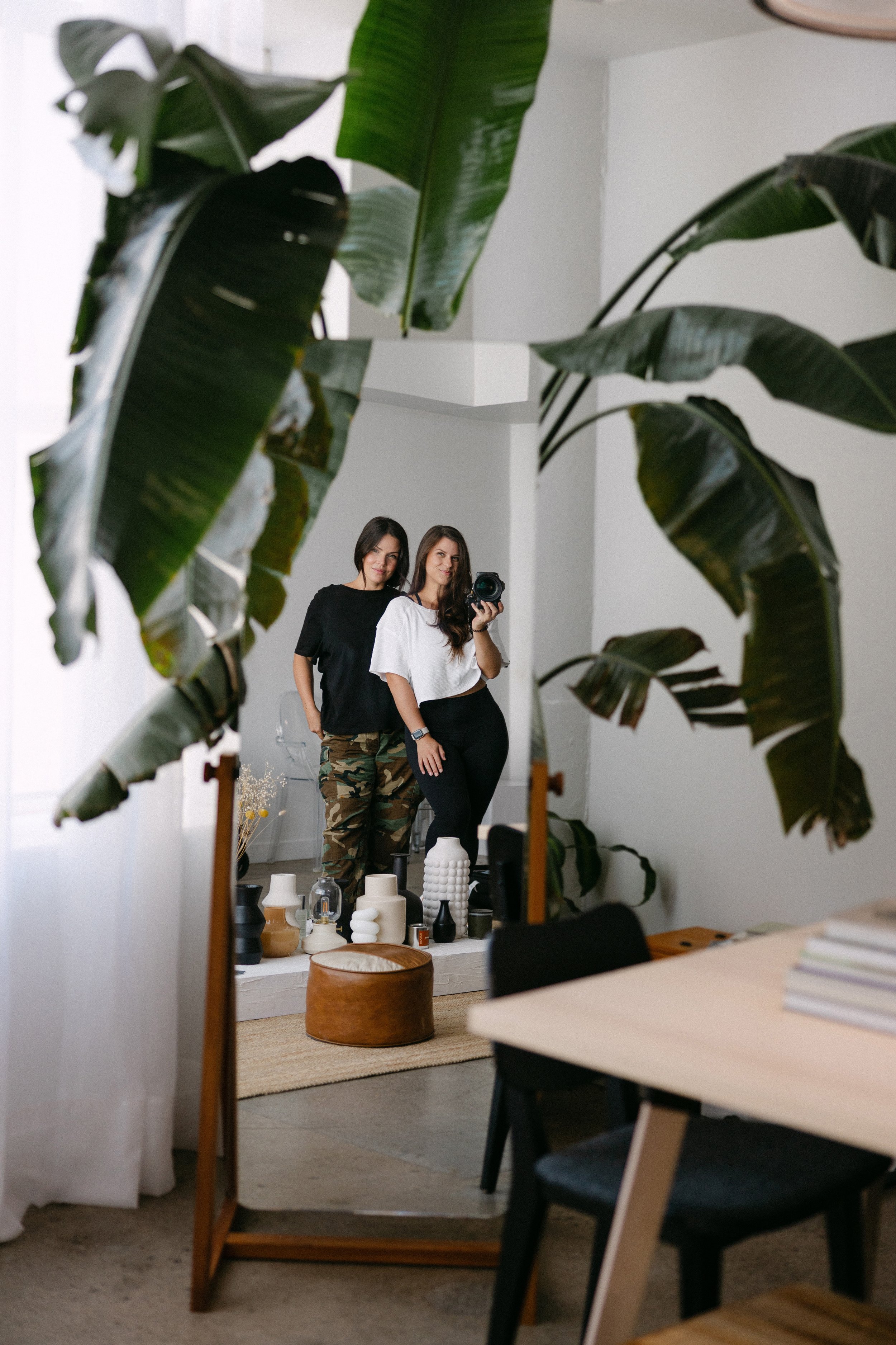 Two women taking a mirror selfie in a room decorated with vases and plants, with a large leafy plant in the foreground.