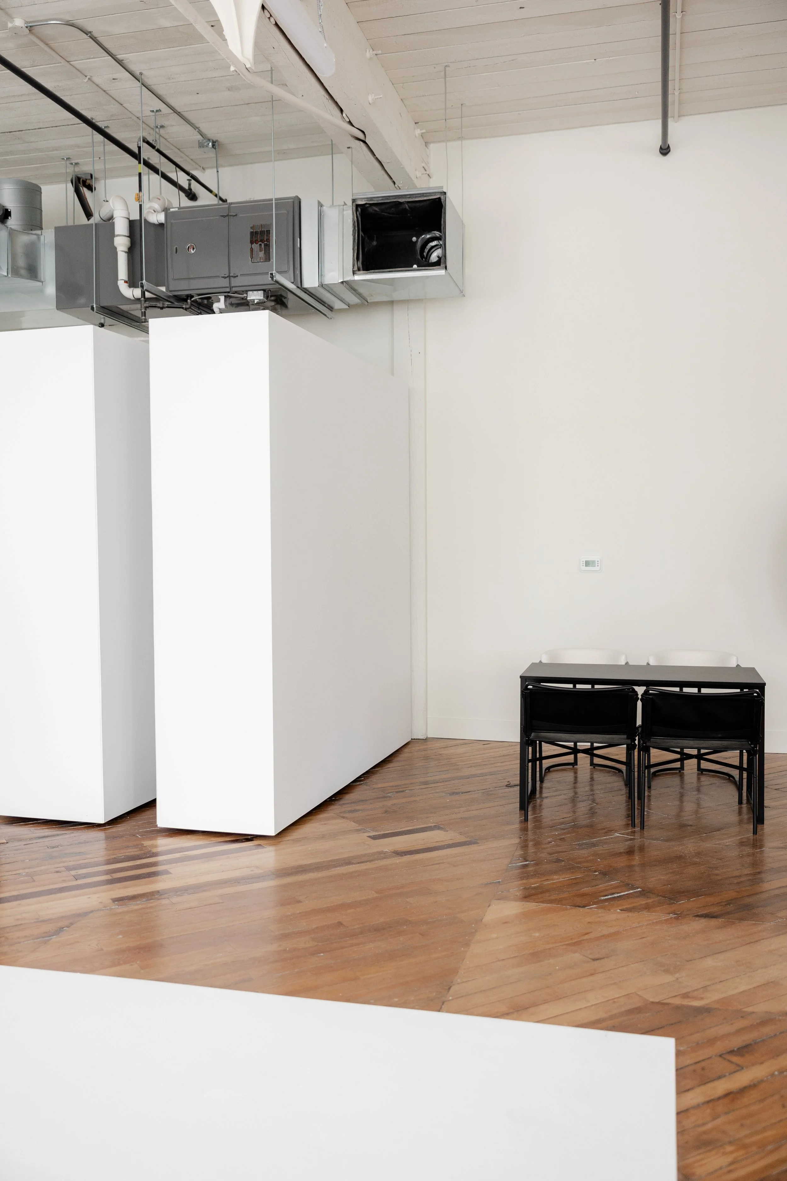An empty room with wooden floors, white walls, and industrial ceiling vents. A small black table with four black chairs is positioned against the wall.