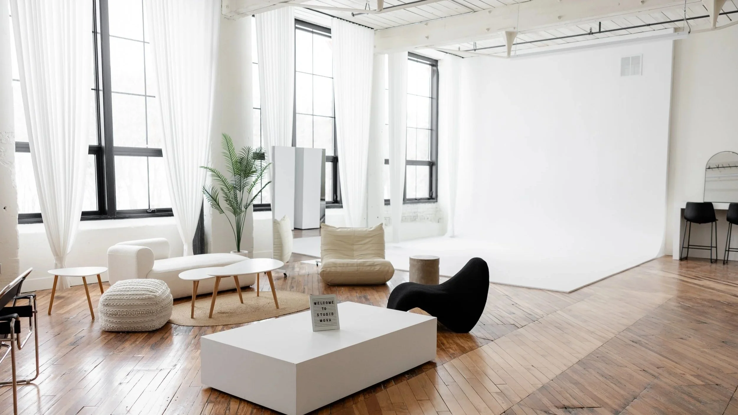Bright studio with large windows, white walls, wooden floors, and modern furniture including sofa, chairs, and a white backdrop for photography or filming.