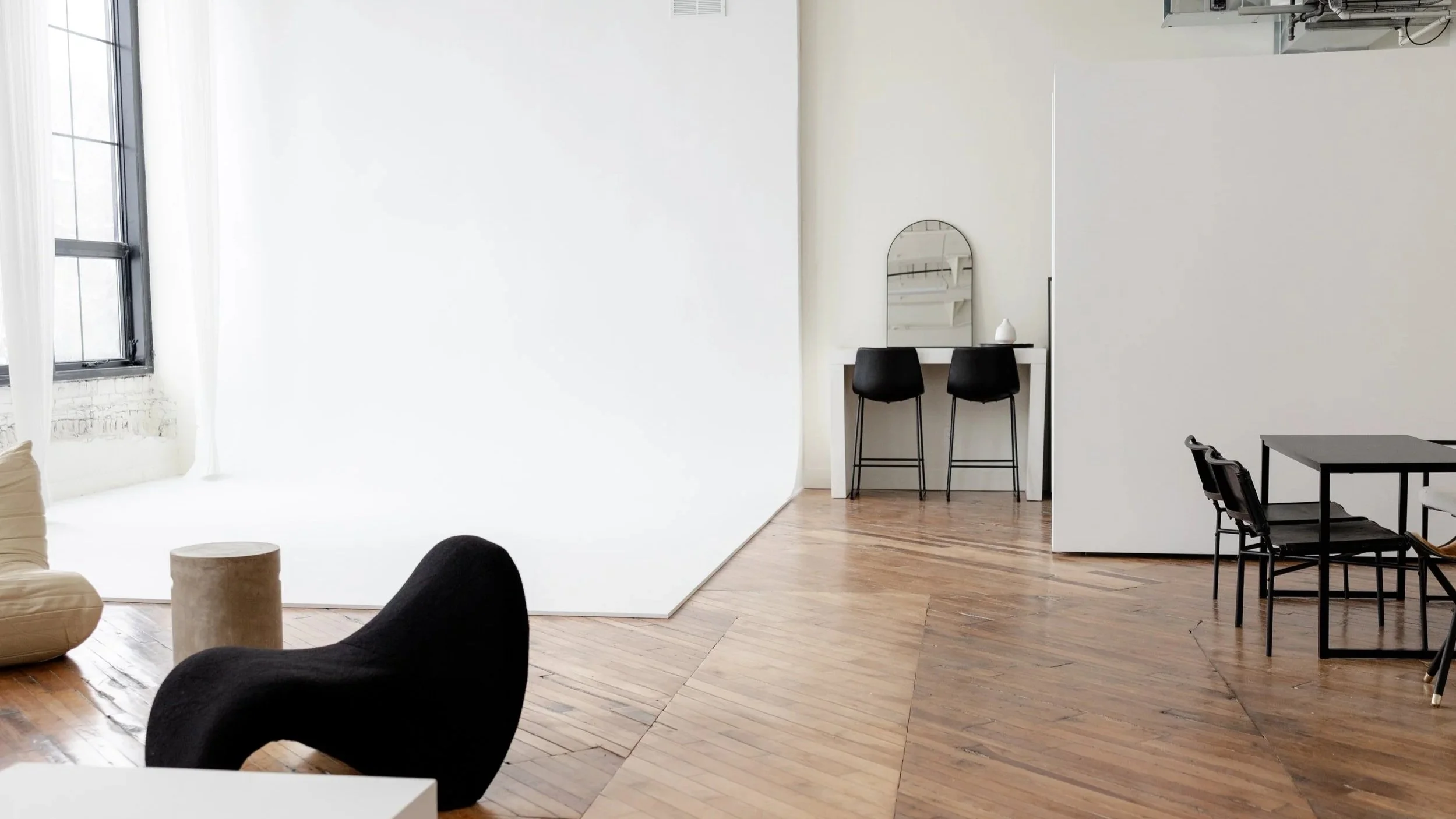 Modern minimalistic living space with wooden flooring, black and beige chairs, white walls, large windows with white curtains, and a white partition wall with a small white table and two black chairs with a mirror.