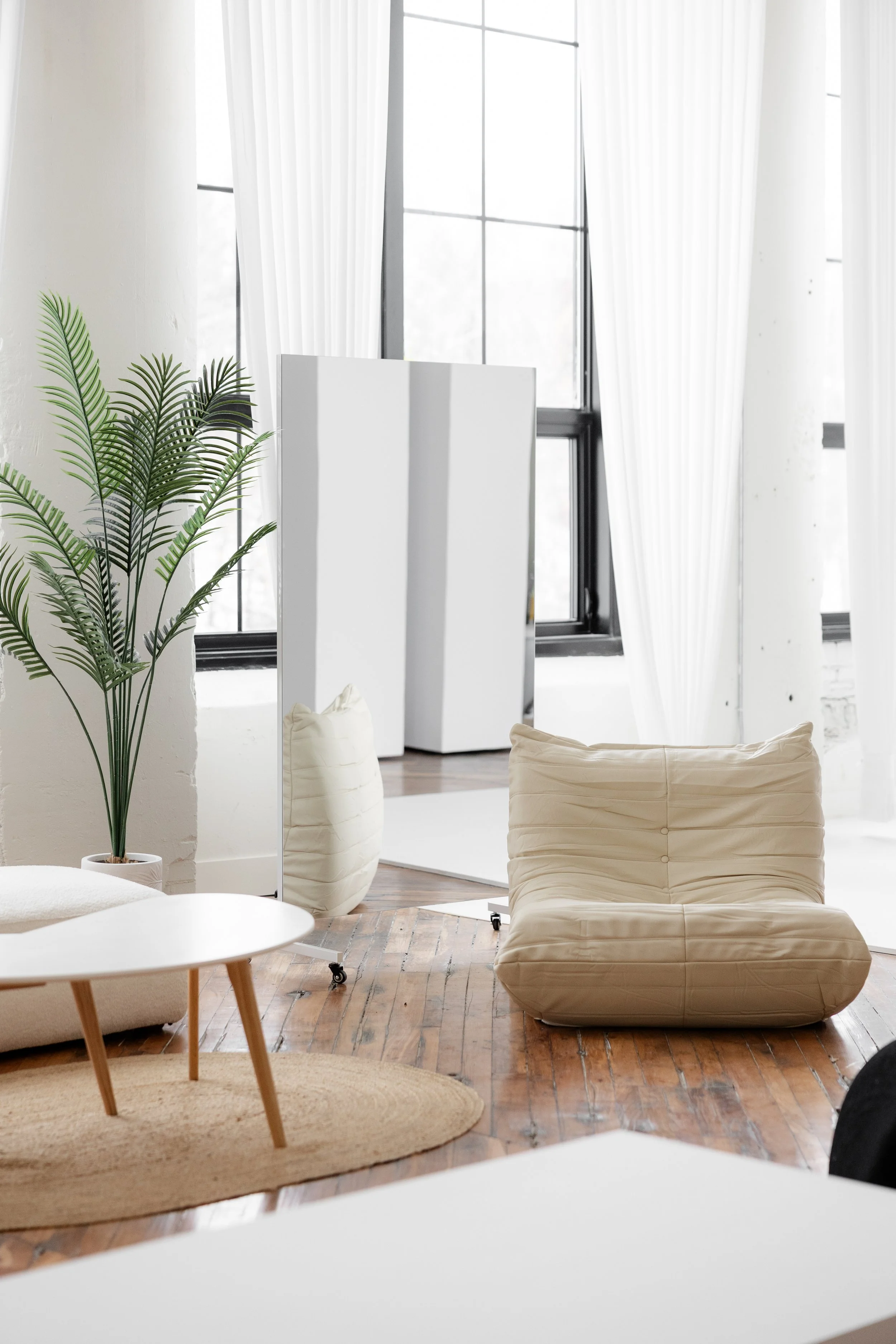 A modern, minimalist living room with large windows, sheer white curtains, a potted plant, a white standing mirror, a beige cushioned lounge chair, a round wooden coffee table on a beige rug, and a white vase on the floor.