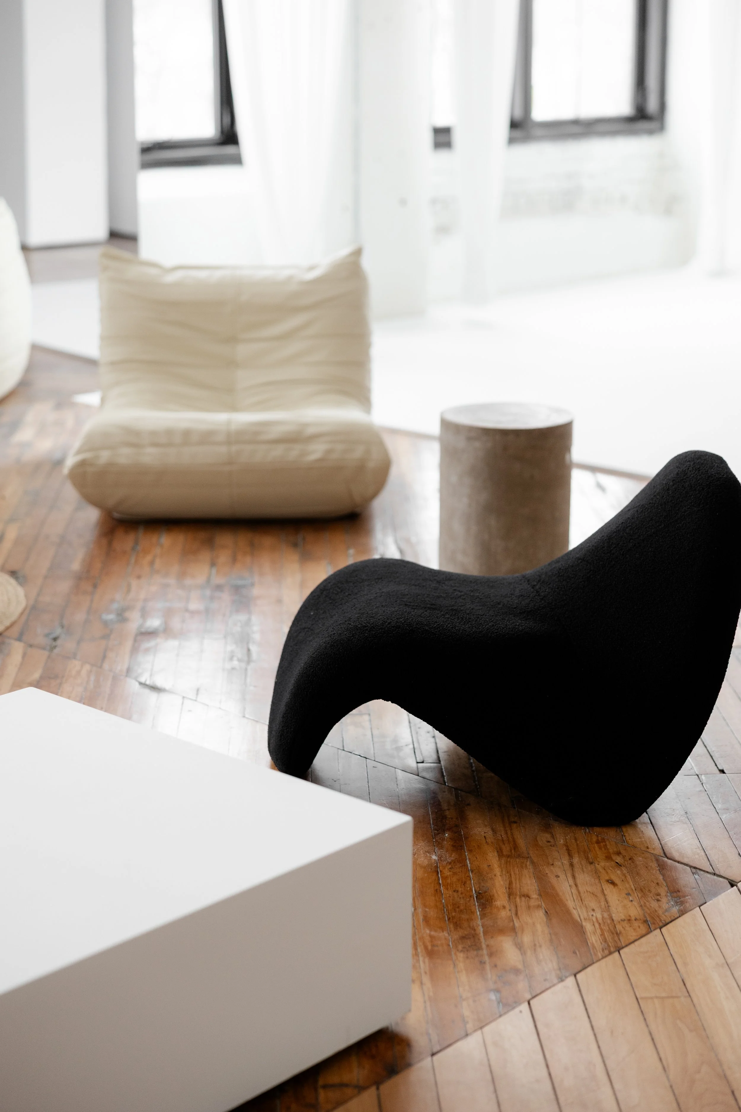 Modern minimalist living room with black sculptural lounge chair, white cushioned chair, concrete cylinder side table, and white coffee table on wooden floor, large windows with white curtains in background.