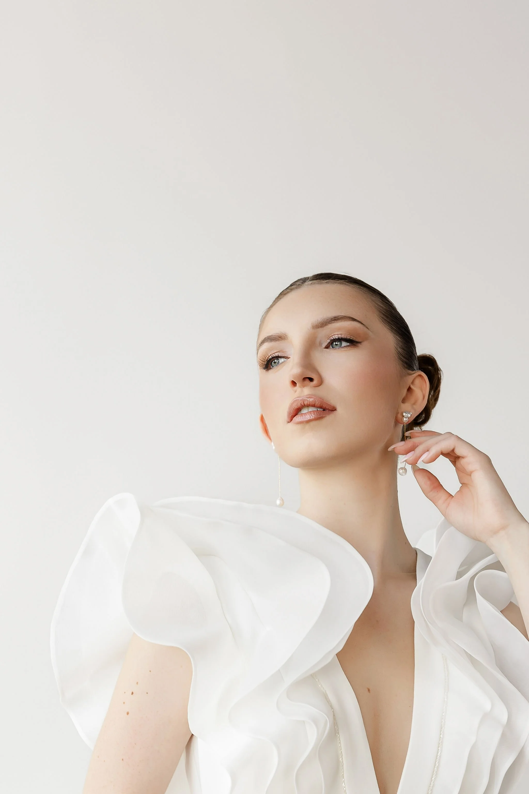 A woman with light skin and blue eyes is wearing a white, ruffled, off-the-shoulder dress and pearl earrings. She has her hair styled in a sleek bun and is posing against a plain white background.