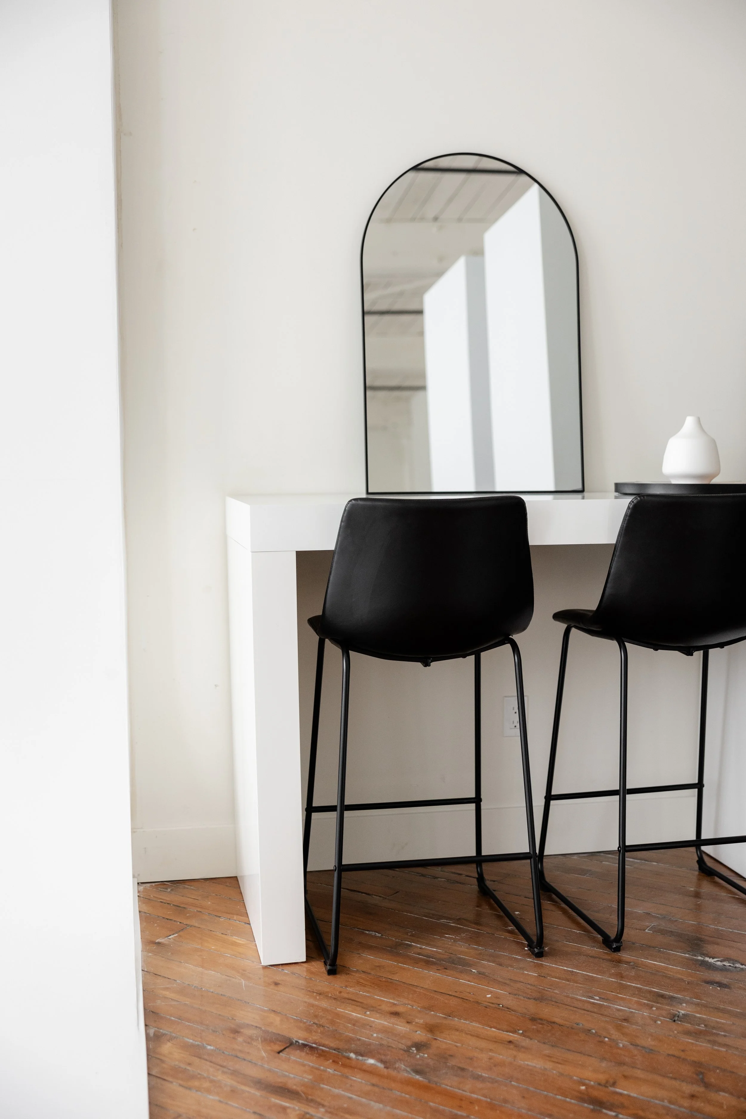 A modern interior space featuring a white countertop with a black-framed mirror above, two black barstools with thin metal legs, a white decorative vase, and a wooden floor.