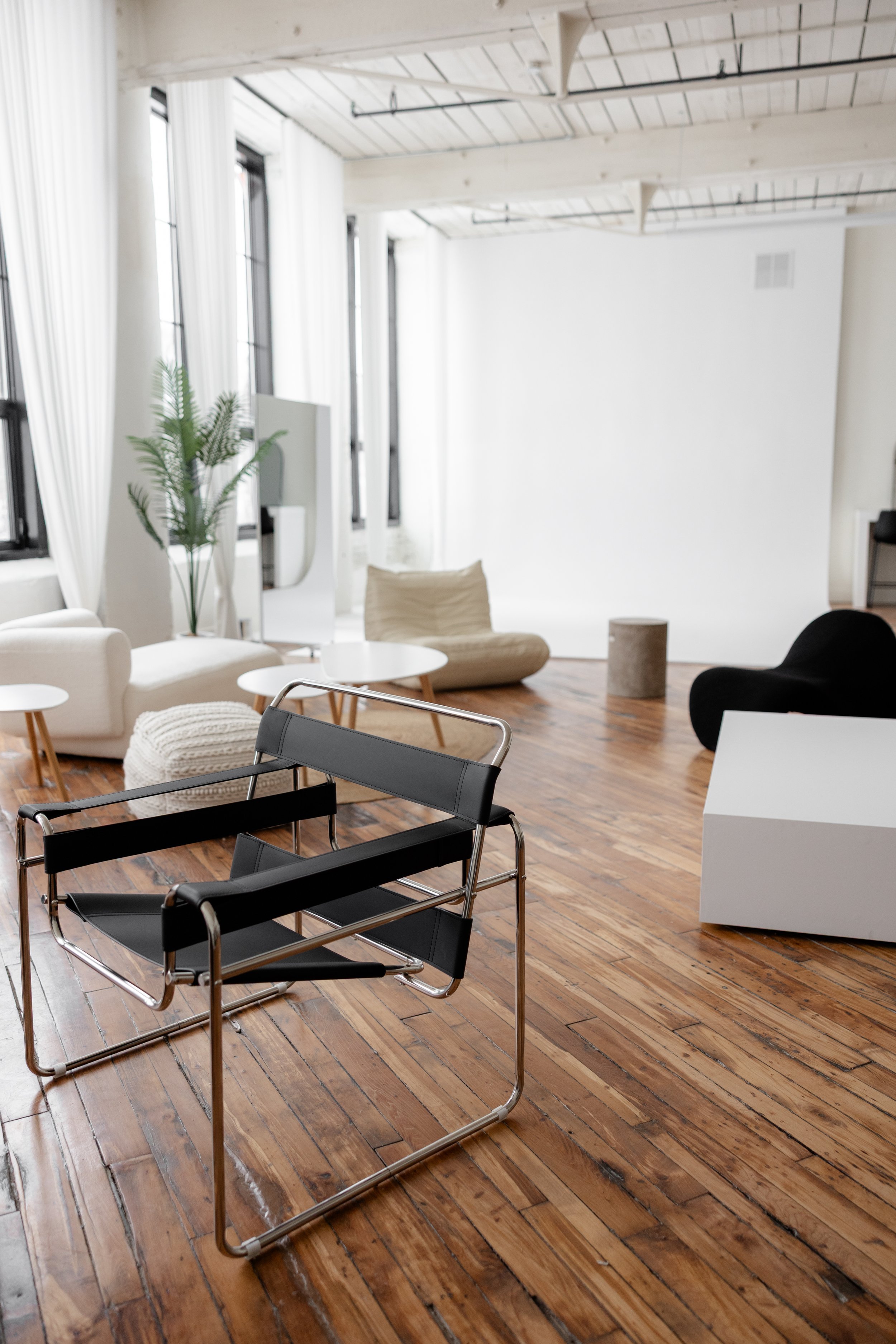 Modern minimalist living room with black lounge chair, white and cream seating, wooden floor, and large windows with white curtains.