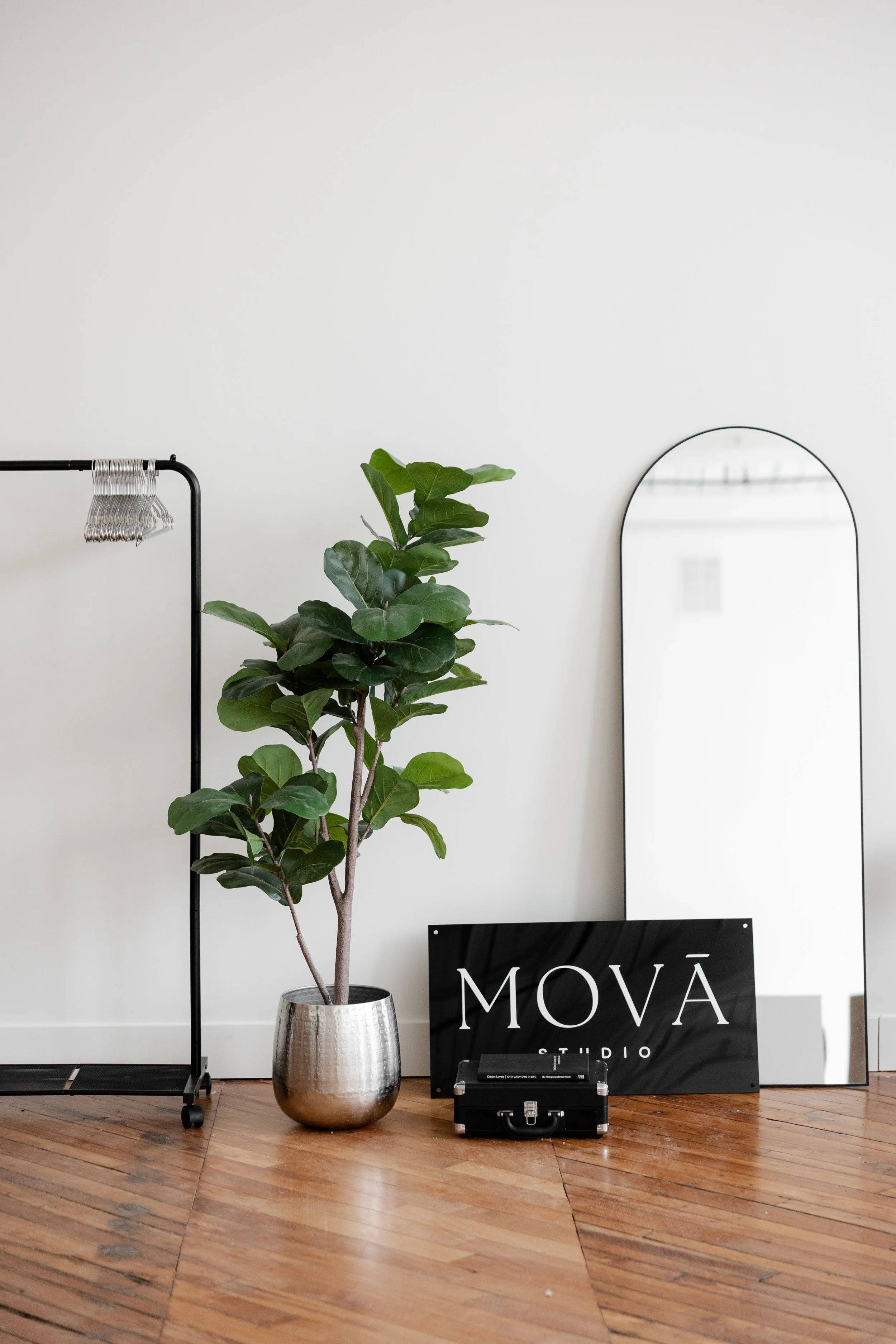 Interior decor scene with a potted green plant, a black sign reading 'MOVA STUDIO', a black briefcase, a tall arched mirror, and a clothing rack with hangers against a white wall and wooden floor.