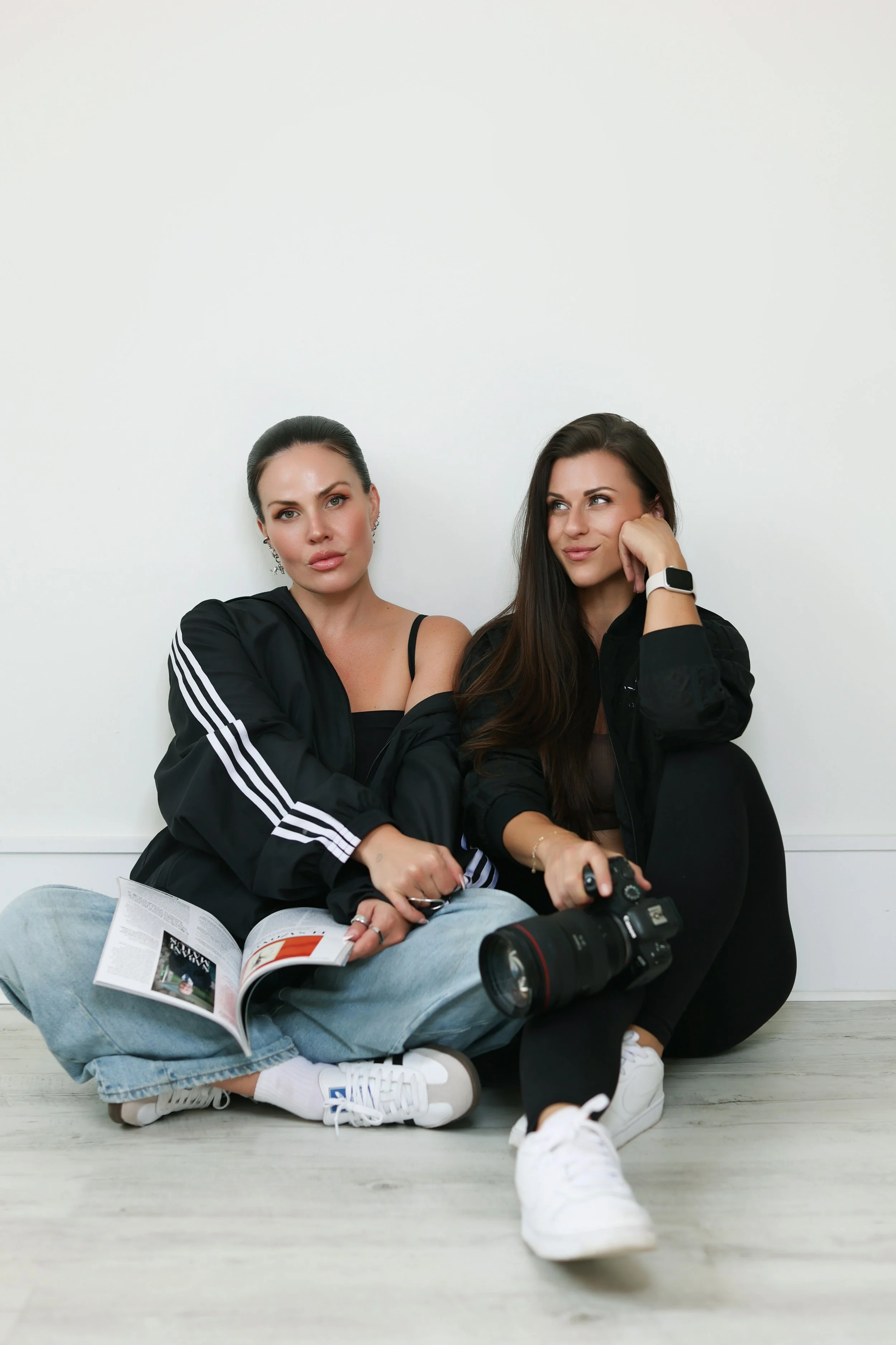Two women sitting against a white wall, one holding a camera and magazine, both wearing black jackets and sneakers.