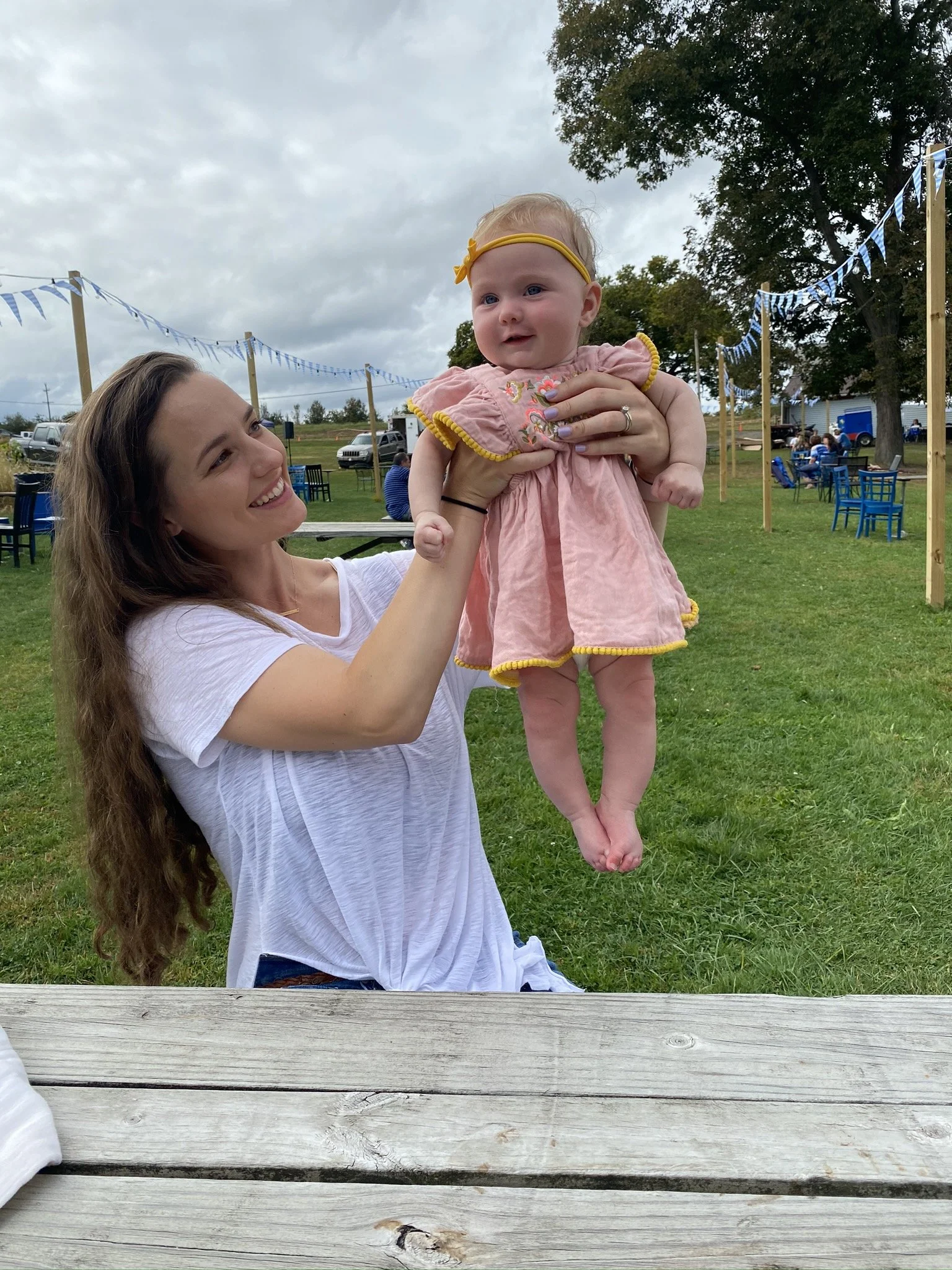 A woman holding a young girl outdoors at a festive event with blue and white decorations. The woman has long brown hair and is smiling while lifting the little girl into the air. The girl is dressed in a pink dress with yellow trim and a yellow headband, and is looking away from the camera.