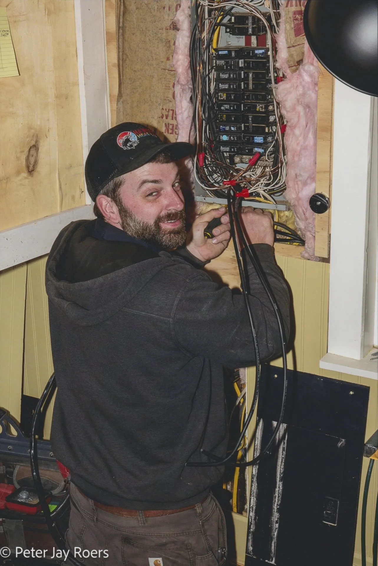 A man with a beard, wearing a black hoodie and a baseball cap, is working on an electrical panel with many circuit breakers and wires. He is smiling and holding a screwdriver.