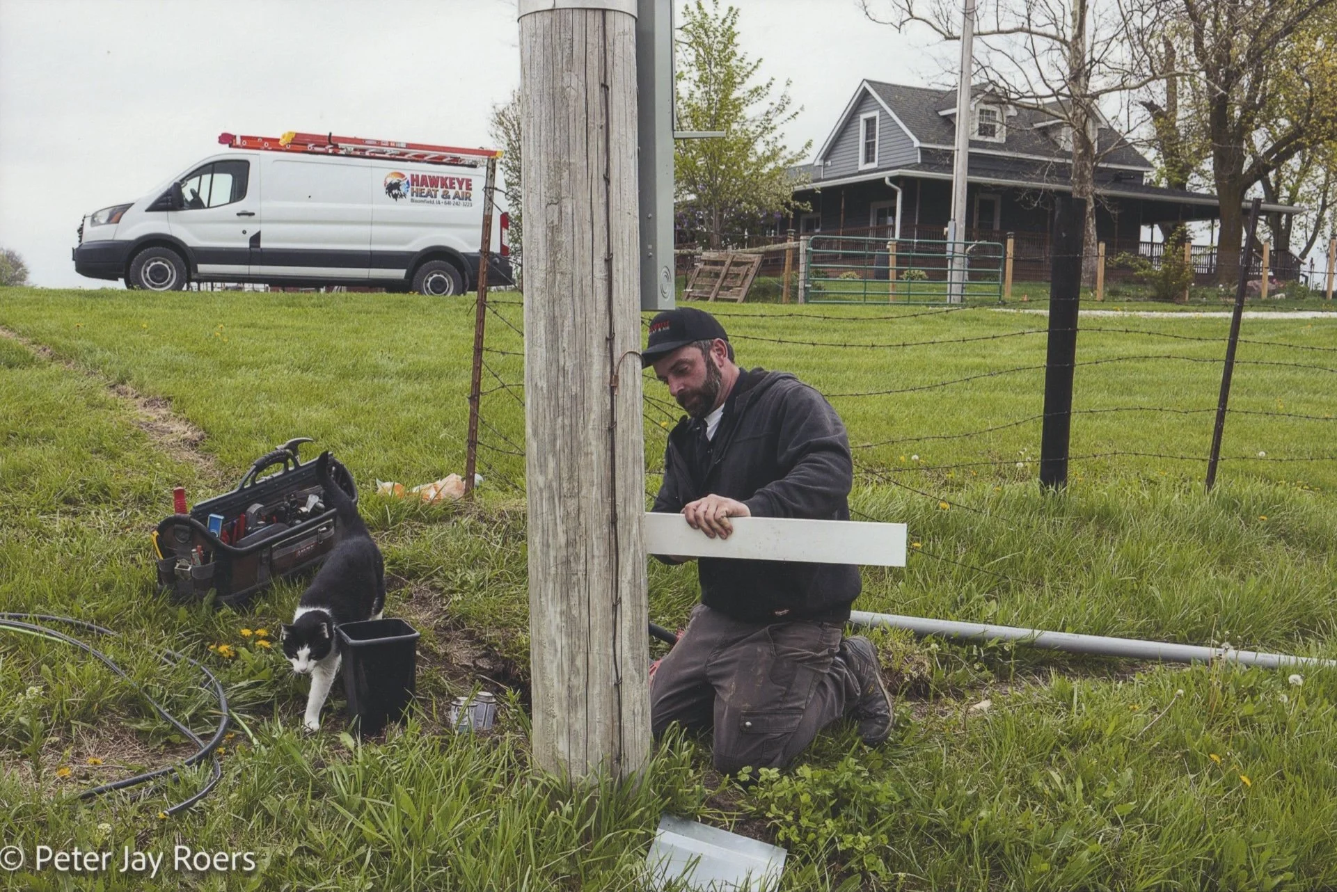 A man kneeling on the grass working on a wooden utility pole during the daytime, with tools nearby and a black and white cat walking in front of him. In the background, a white van with a ladder on top and a house with a porch are visible.