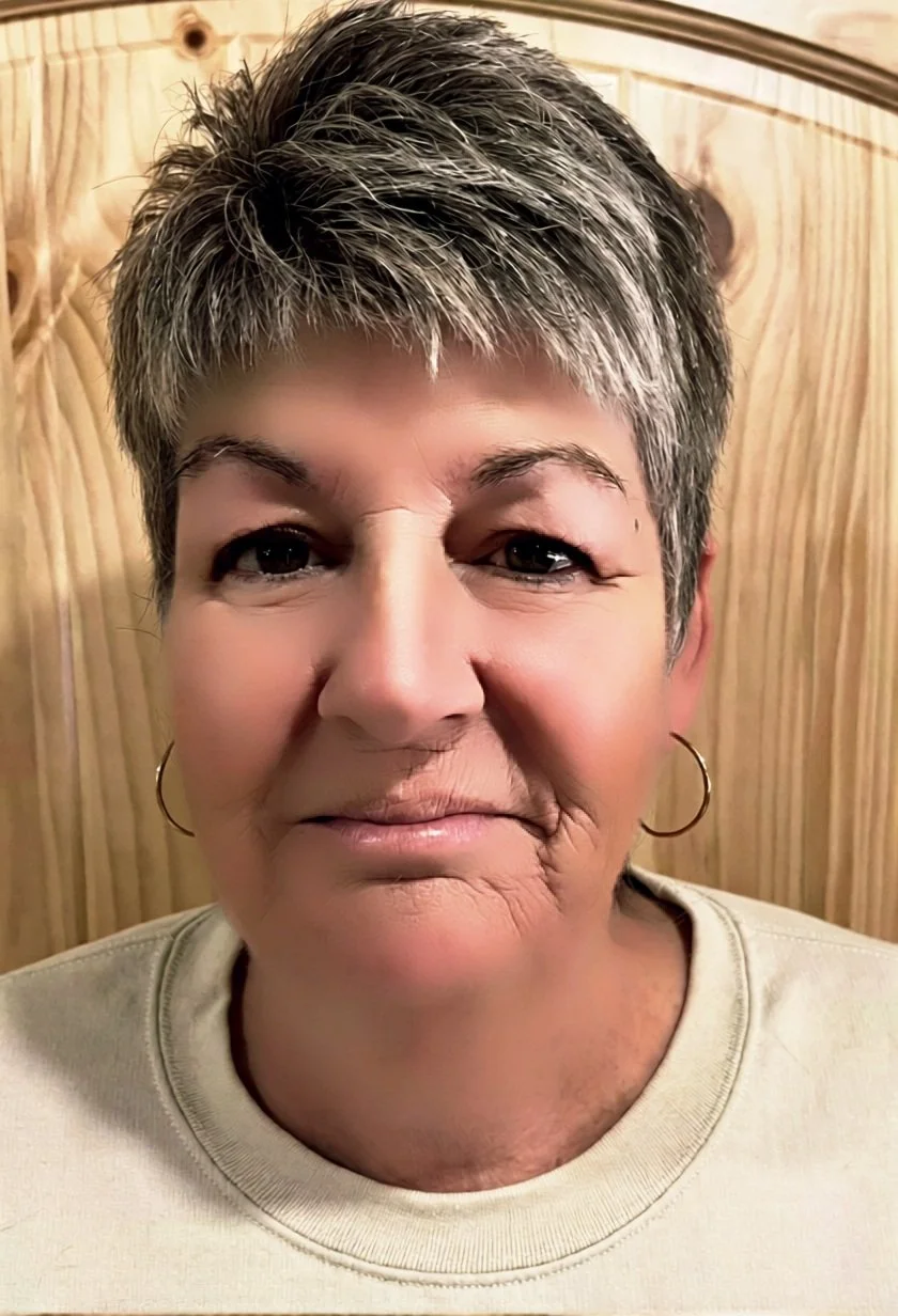 Close-up of a woman with short gray hair, wearing hoop earrings and a light-colored shirt, standing indoors in front of a wooden background.
