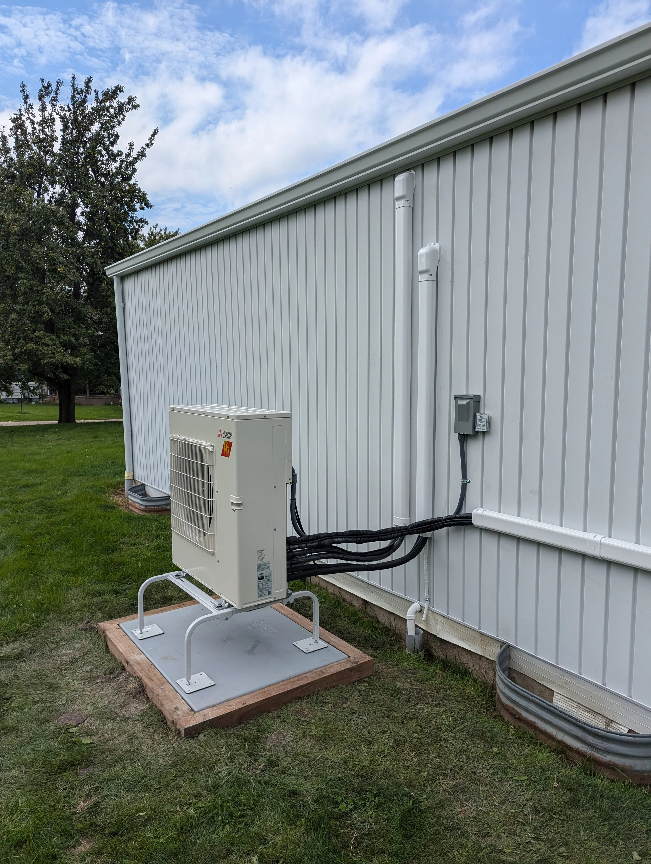 Outdoor HVAC unit installed beside a white building on a grassy area under a partly cloudy sky.