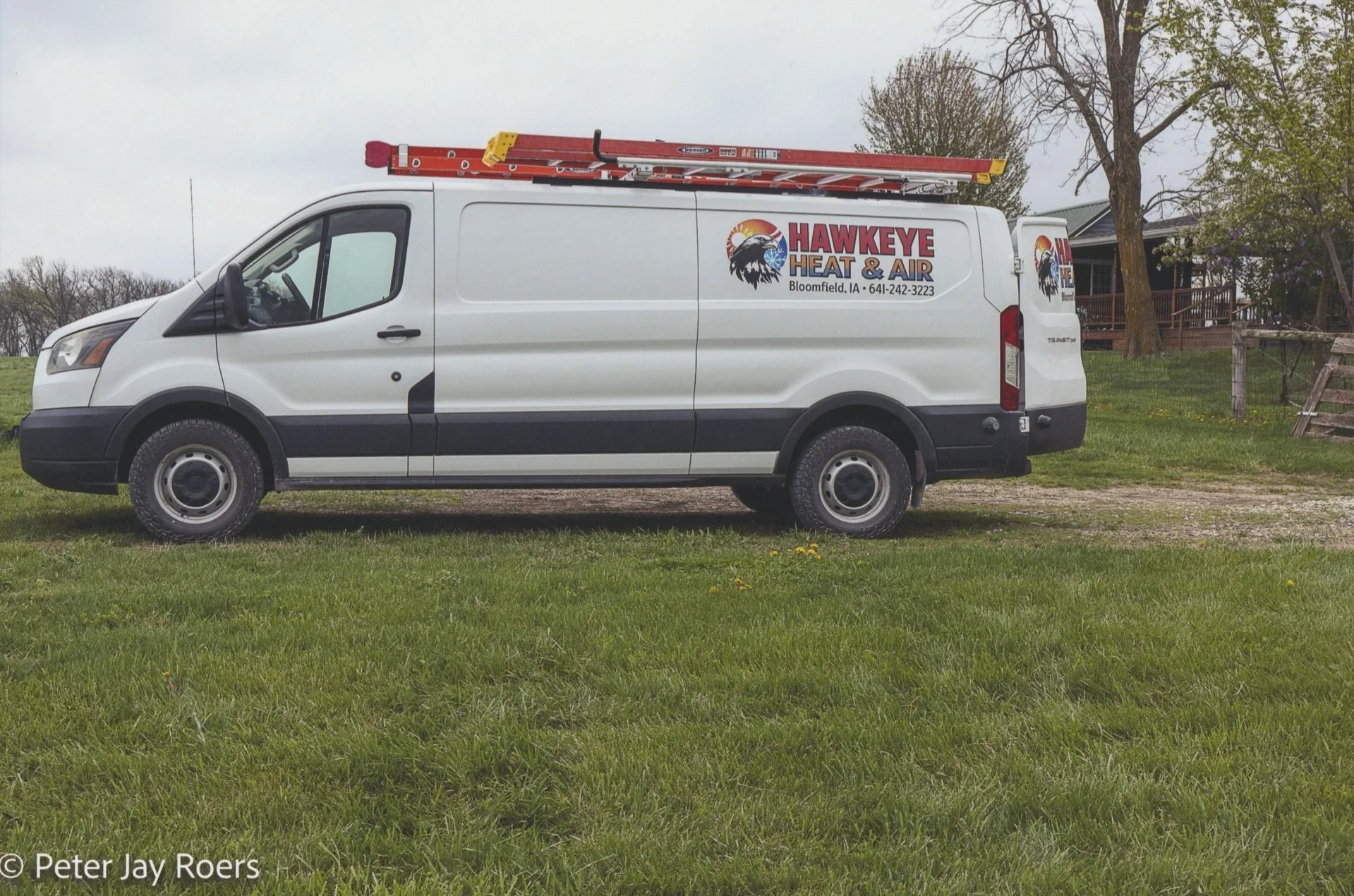A white service van with the logo for Hawkeye Heat & Air parked on a grassy area near a house, with ladders mounted on top.