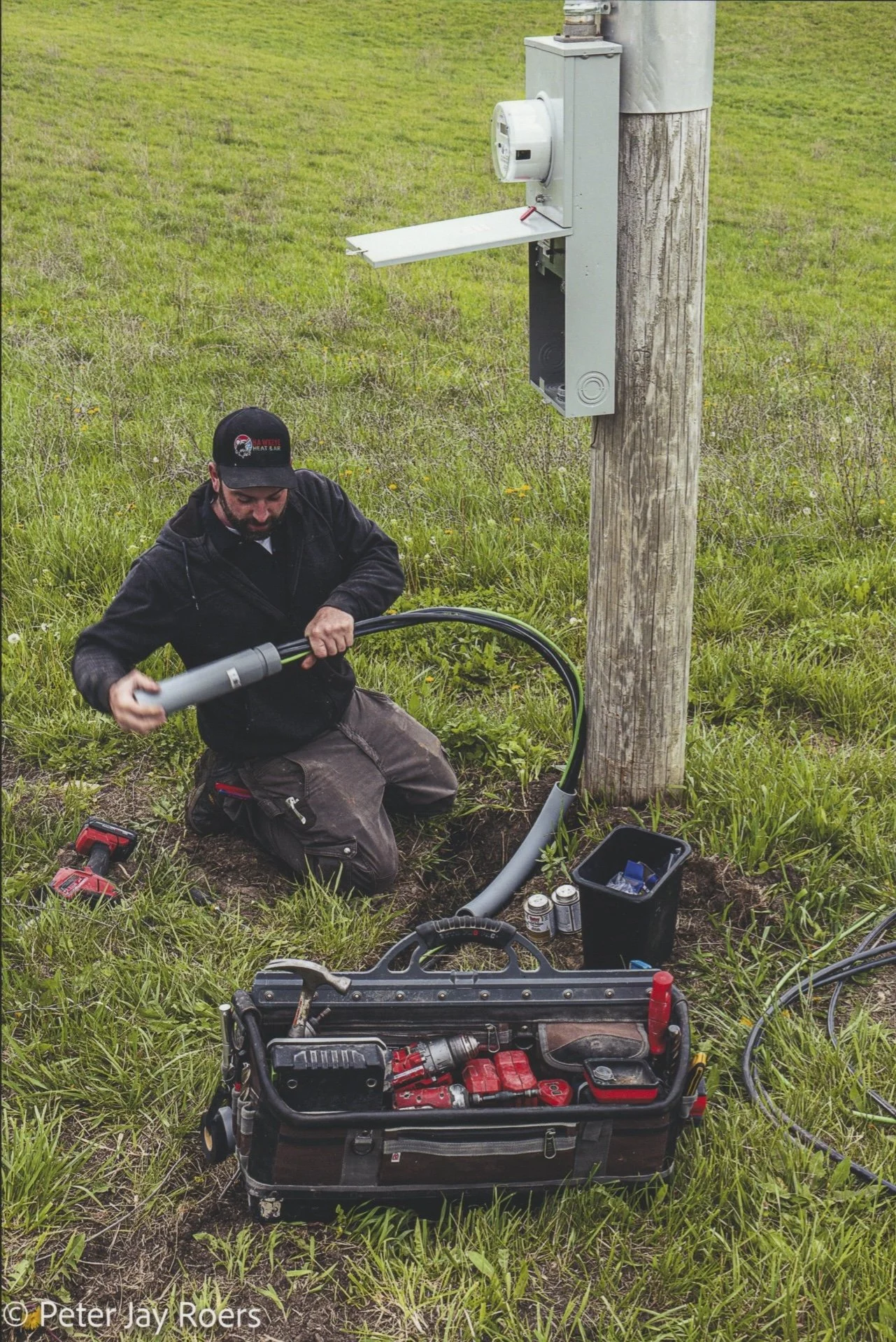 A man kneeling on grass working on an underground utility cable connected to a utility pole, with tools and equipment nearby.