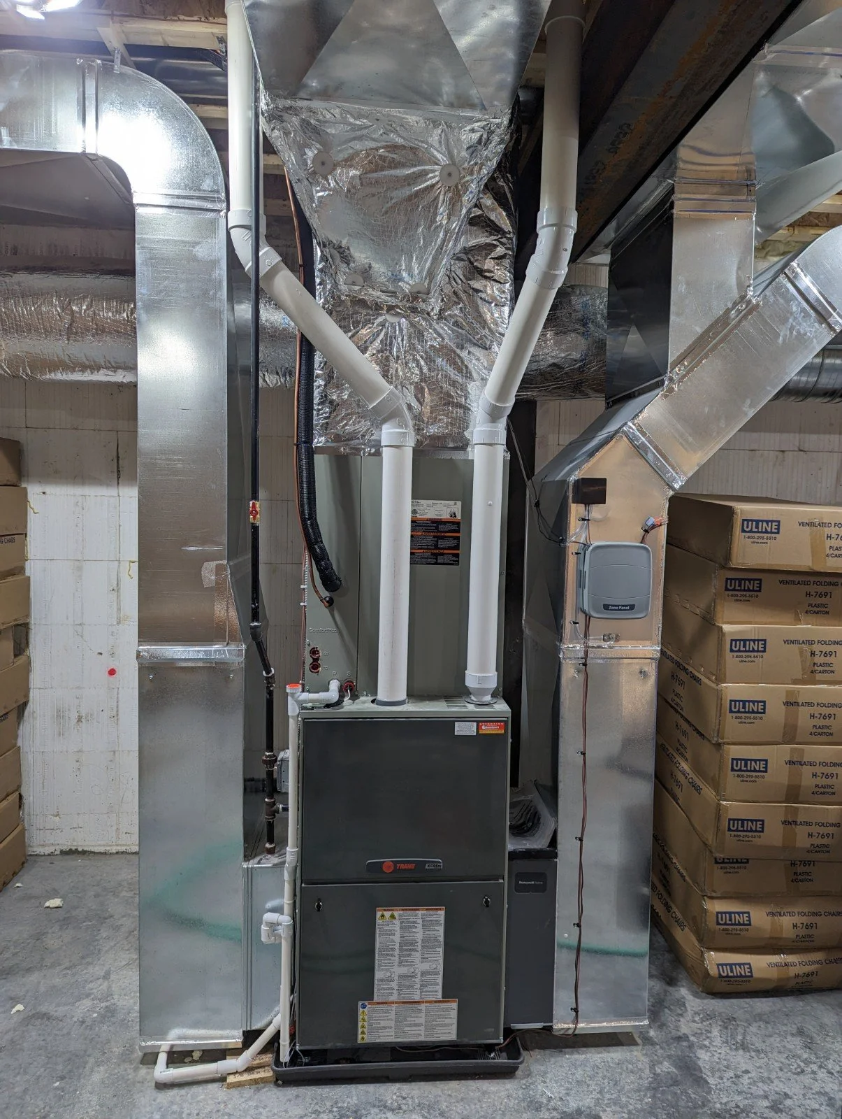 HVAC furnace system with ducts and pipes in a basement, surrounded by boxed packages of Uline storage items.