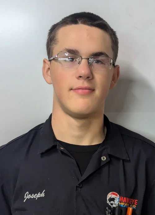 A young man with short dark hair and glasses, wearing a black uniform shirt with a name tag that says Joseph and a logo for Hawkeye Heat & Air.