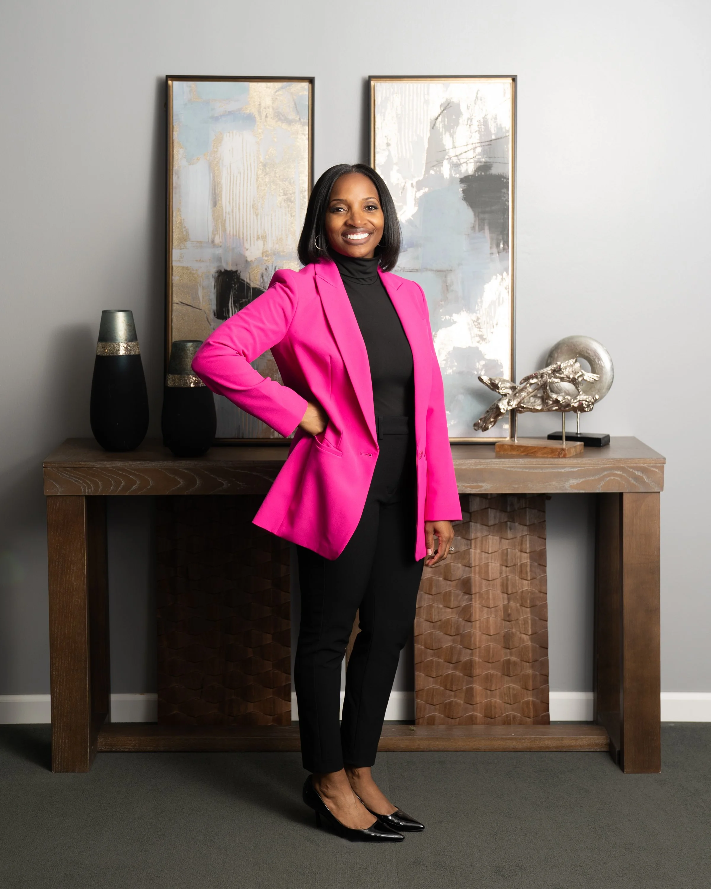 A woman in a pink blazer and black pants standing in front of a wooden console table with abstract artwork and decorative objects, smiling at the camera.