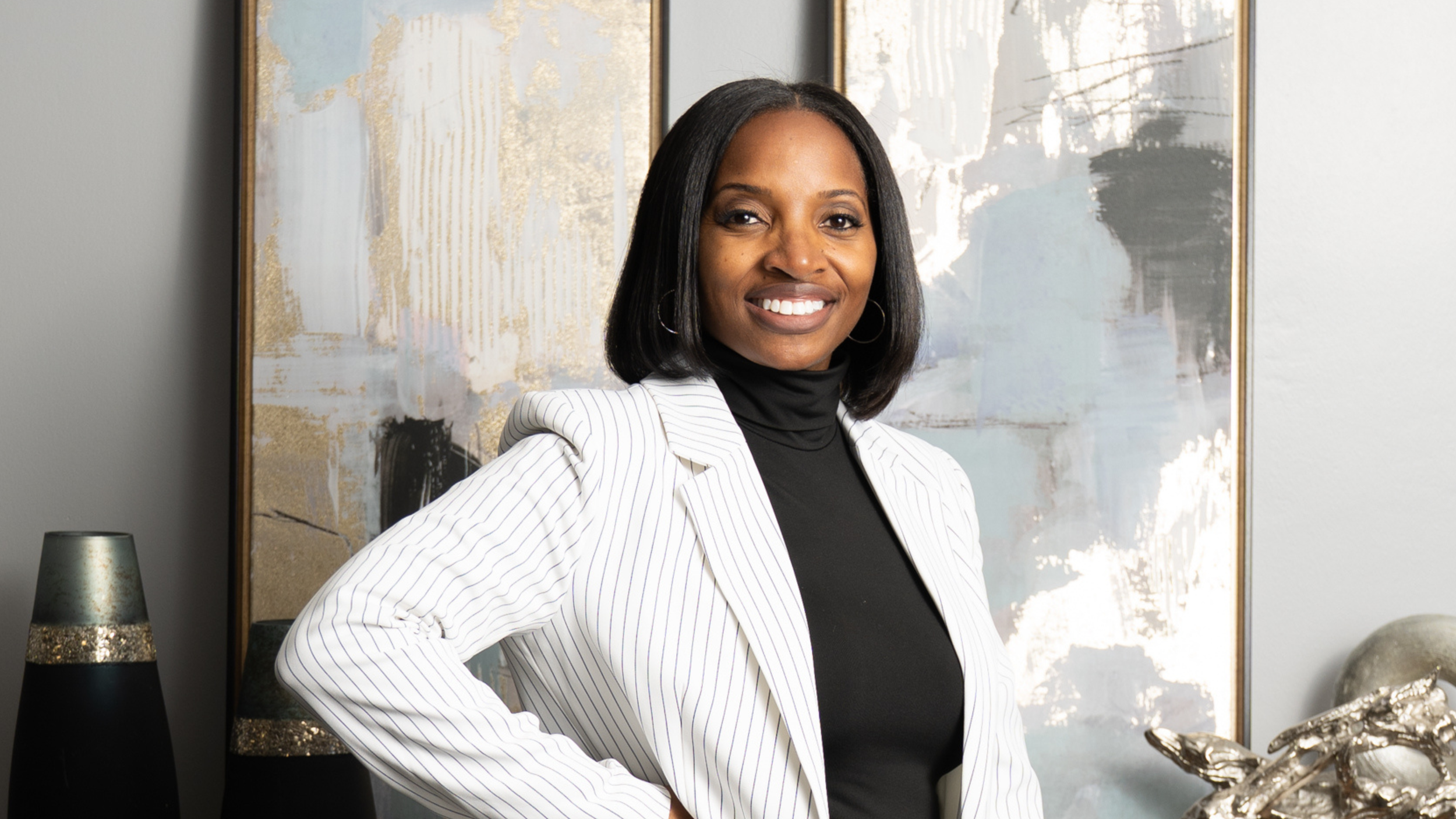 A woman standing in front of abstract artwork with gold, gray, and white tones, smiling with her hand on her hip, wearing a black turtleneck and a white striped blazer.