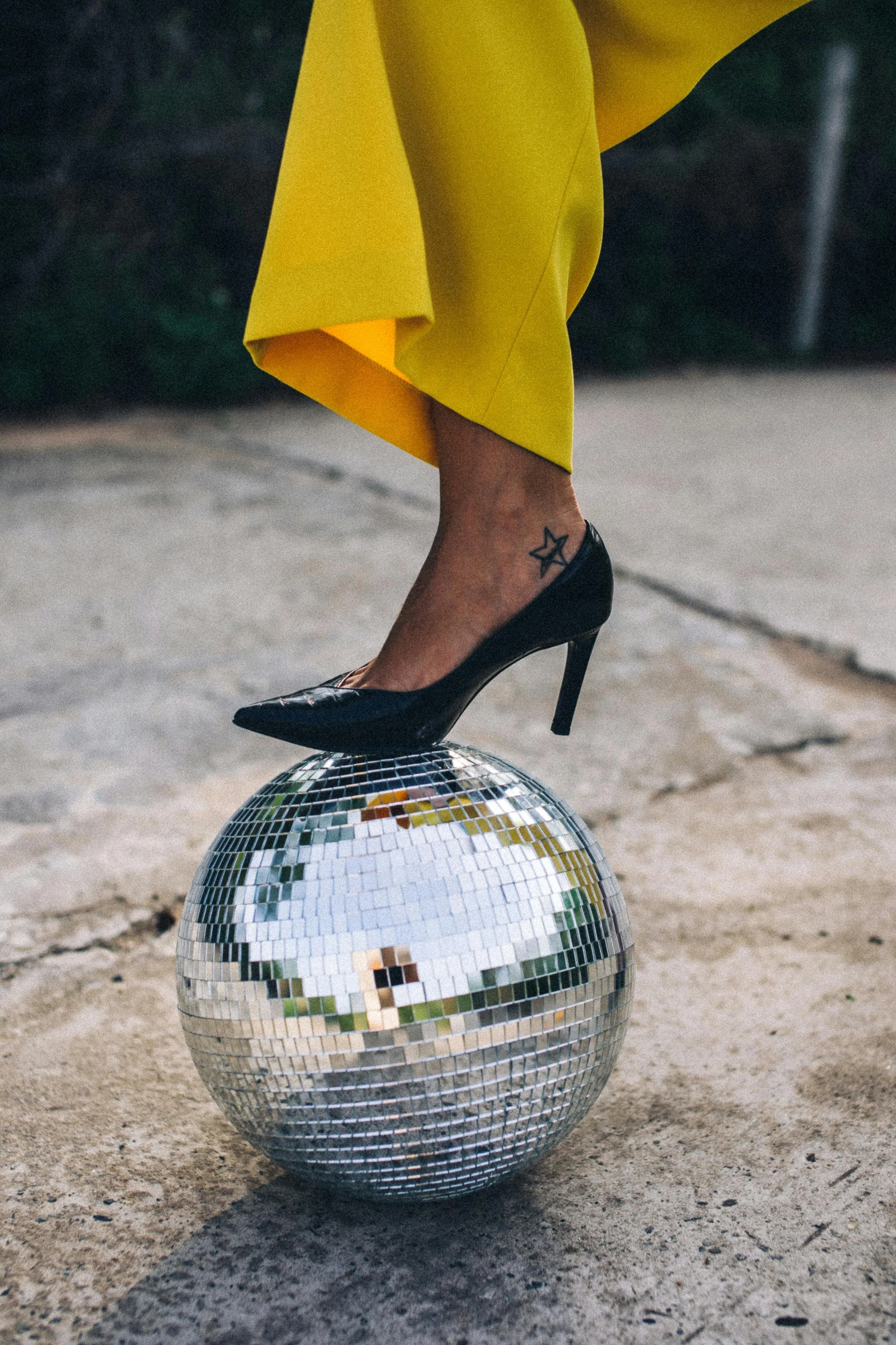 Woman's leg and foot in black stiletto resting on top of silver disco ball that is outside.
