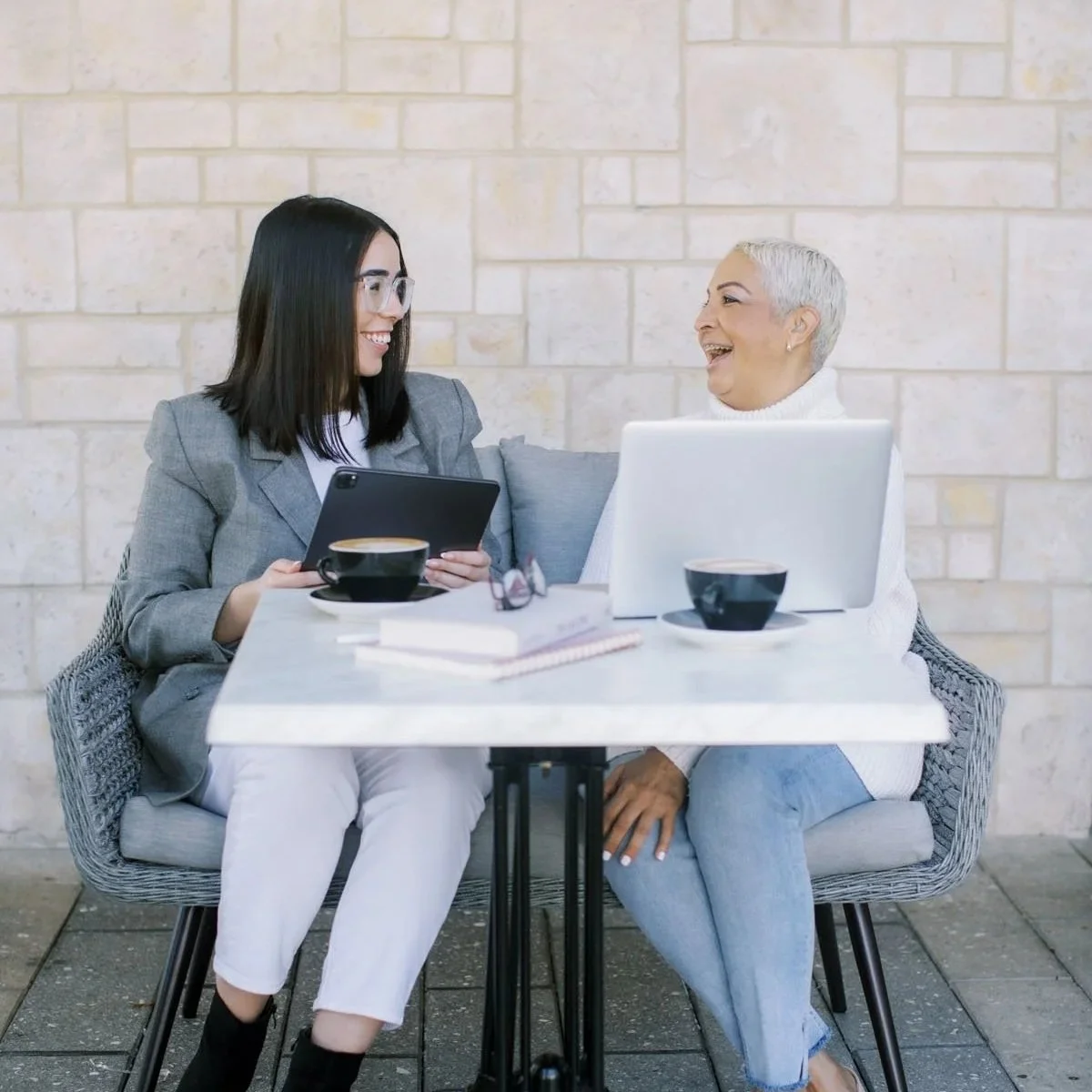 Two women, one younger with shoulder length dark hair and one older with cropped silver hair sitting at an outdoor cafe table with a laptop and tablet and coffees chatting and smiling