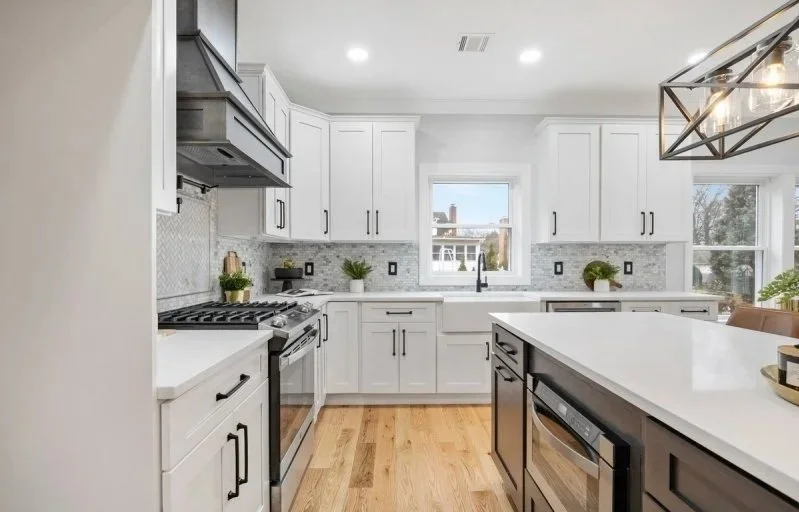 Modern white kitchen with wooden floors, black hardware, and a large island with a microwave, pendant lighting, and windows overlooking a neighborhood.