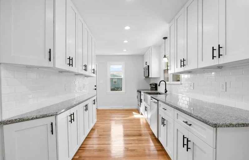 Modern white kitchen with granite countertops, white cabinets with black handles, a window, appliances, and hardwood flooring.