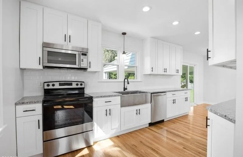 Modern white kitchen with stainless steel appliances, granite countertops, and hardwood floors, featuring a large window above the sink.