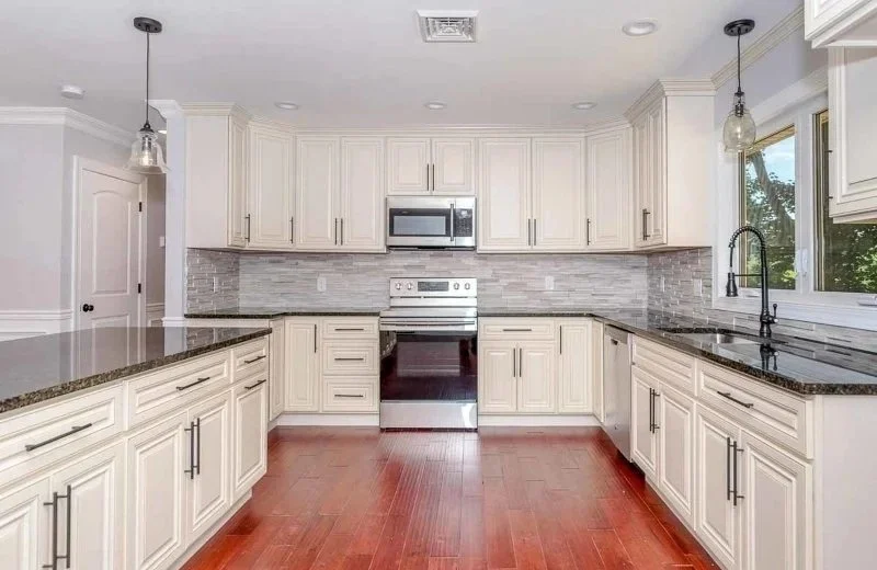 Modern kitchen with white cabinets, dark granite countertops, gray tile backsplash, stainless steel appliances, and hardwood flooring.