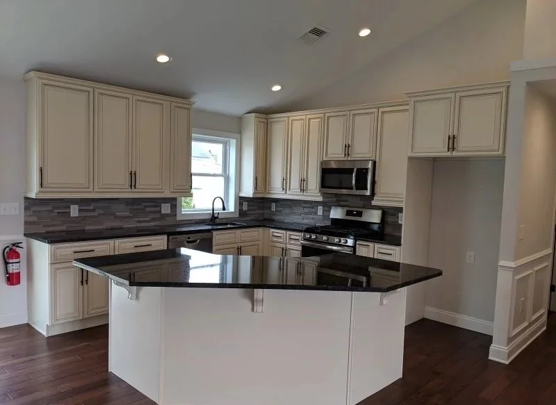 Modern kitchen with white cabinets, black countertops, stainless steel appliances, gray tile backsplash, and a central island with a black top. There is a window above the sink and recessed lighting.
