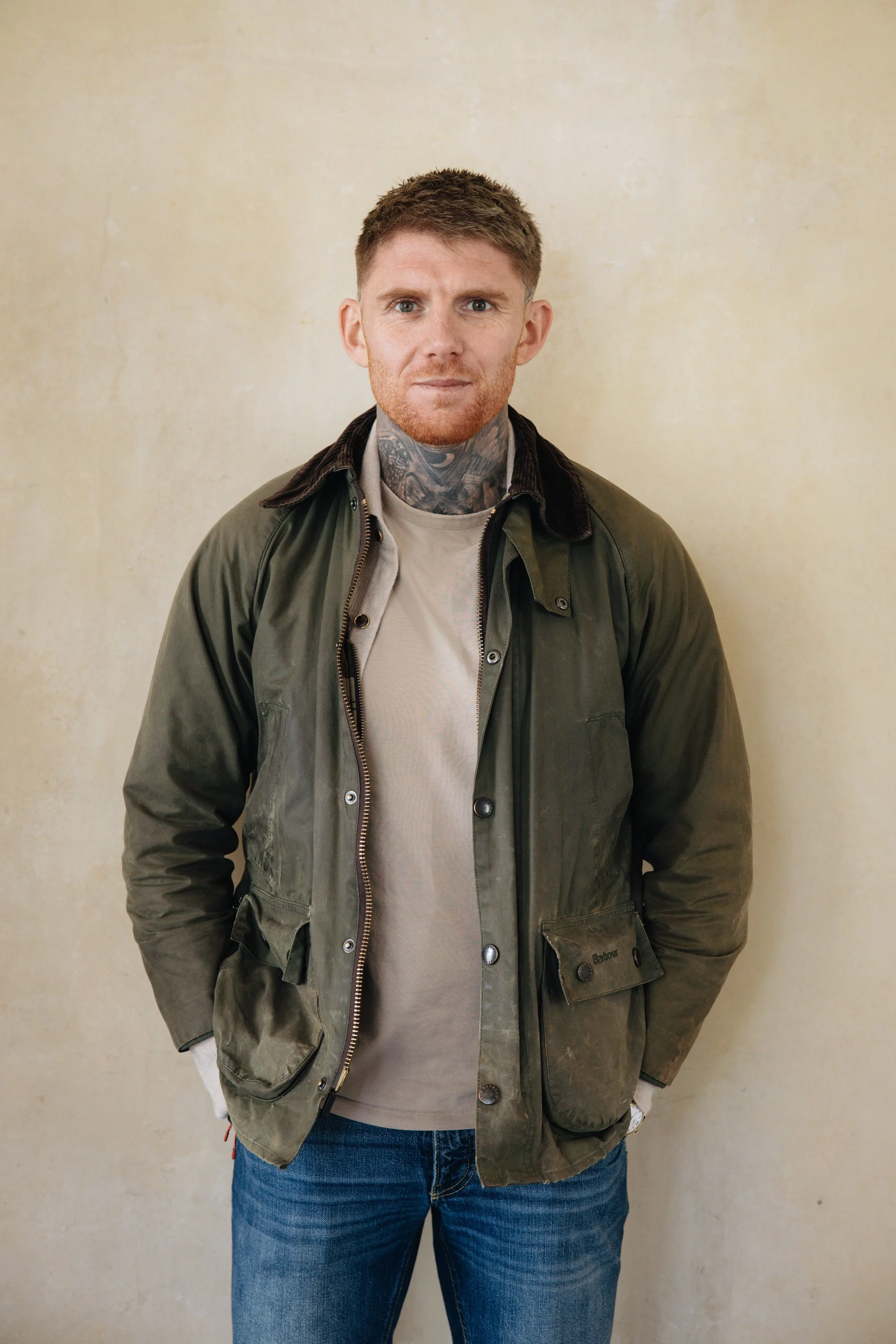 A man with red hair, a beard, and tattoos on his neck, wearing a green jacket over a beige shirt and blue jeans, standing against a plain beige wall.