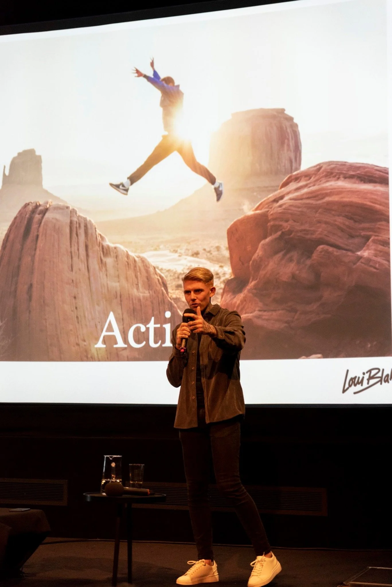A young man in a brown jacket and black pants holding a microphone stands in front of a large screen showing an image of a person jumping between rocks in a desert landscape at sunset, with the word 'Acting' visible at the bottom.