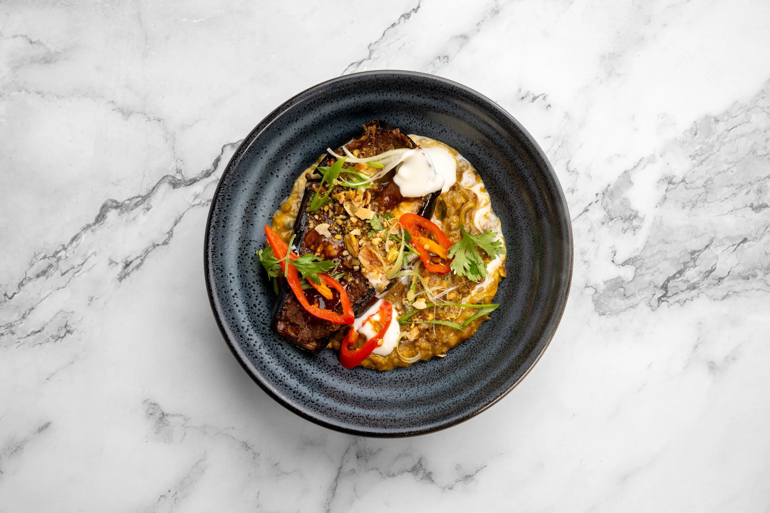 A black plate with a serving of curry topped with grilled eggplant, red chili slices, chopped nuts, cilantro, green onions, and dollops of creamy white sauce, placed on a white marble surface.