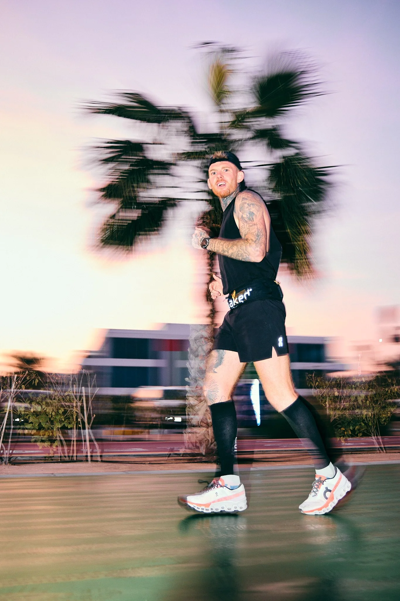 A tattooed man running outdoors with a palm tree in the background, pink and purple sunset sky, wearing black sportswear, white sneakers, and black knee-high compression socks.