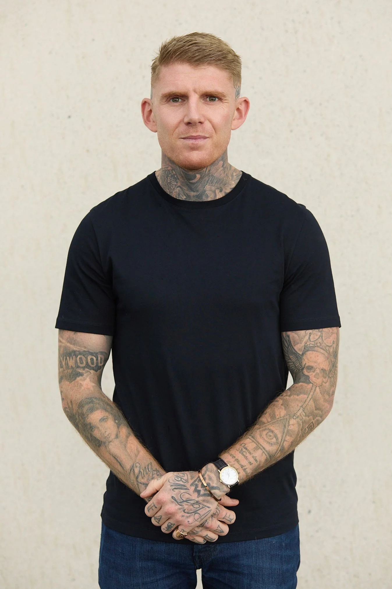 A man with blond hair and tattoos on his neck and arms stands against a plain, off-white wall, wearing a black t-shirt and blue jeans.