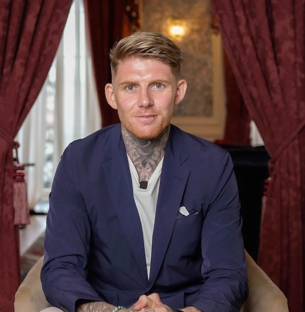 A man with styled blonde hair, light skin, and tattoos on his neck and arms, wearing a navy blazer over a white shirt, sitting in a room with red curtains and warm lighting.